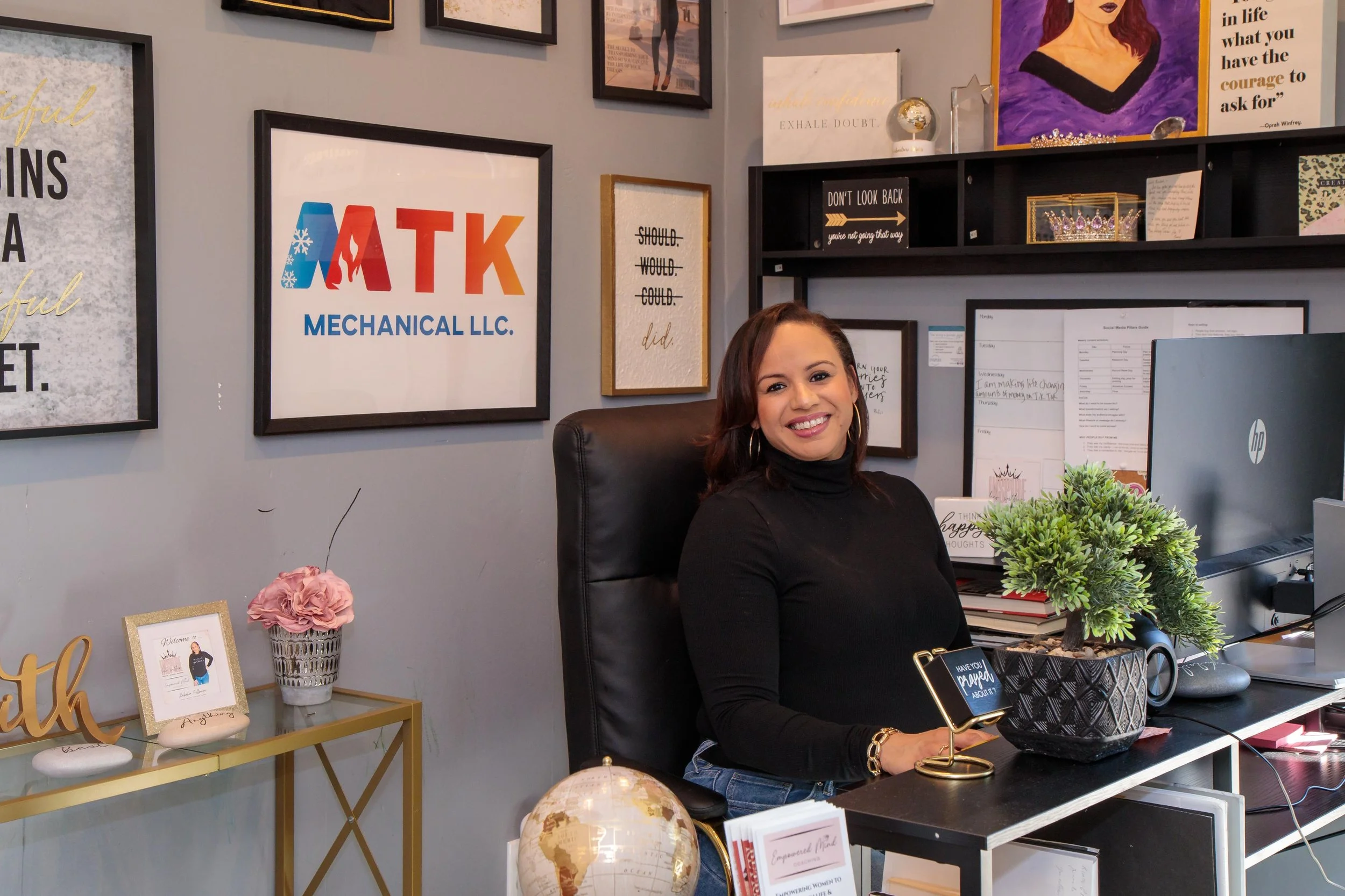 A smiling woman sitting at a desk in an office, surrounded by framed inspirational quotes and artwork on the walls, with a computer monitor in front of her and potted plants on the desk.