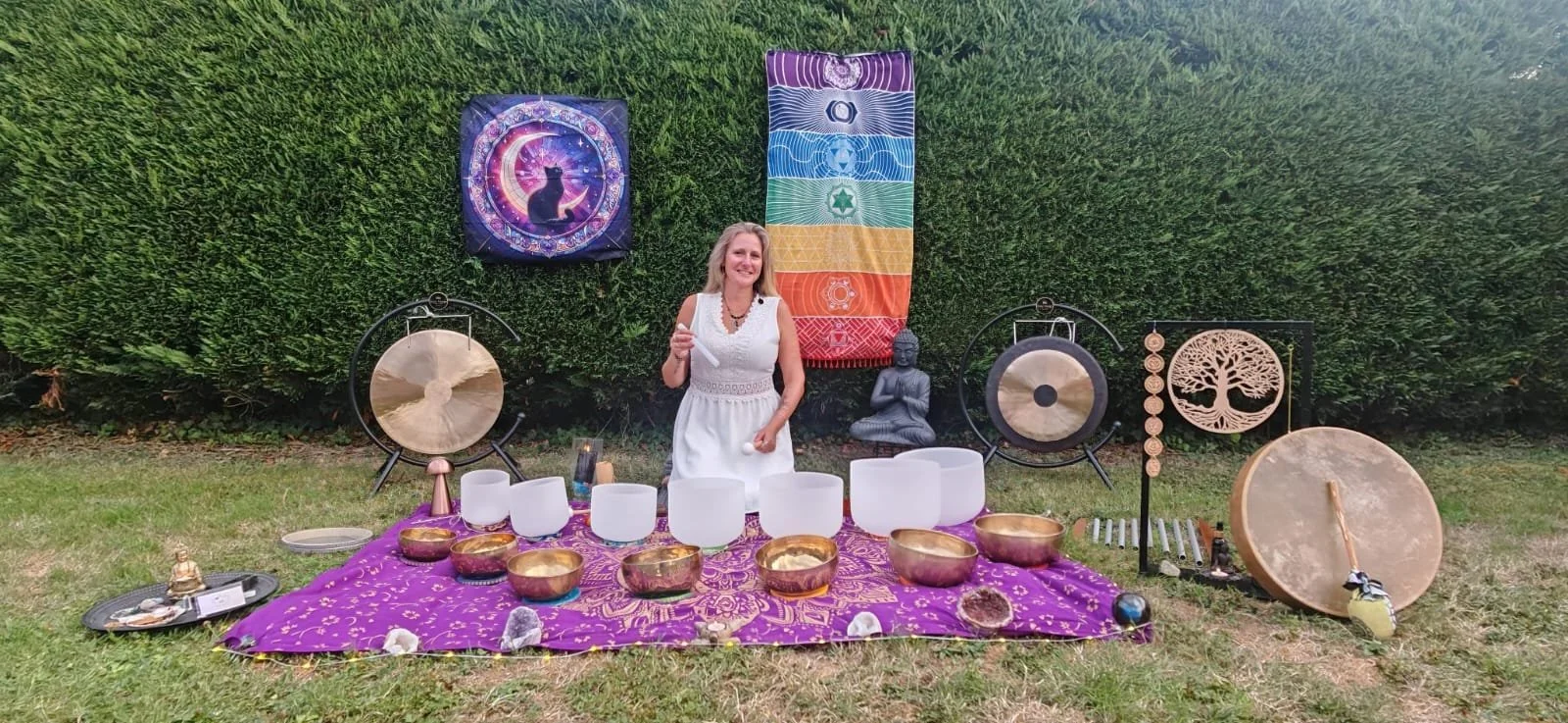 Une femme souriante se tient devant une table de cristal avec des bols en métal, des bols de cristal blanc, et diverses boîtes et objets, entourée de décorations spirituelles y compris des gongs, un tapis violet avec des motifs or, et des sculptures, dans un espace extérieur avec buissons verts.