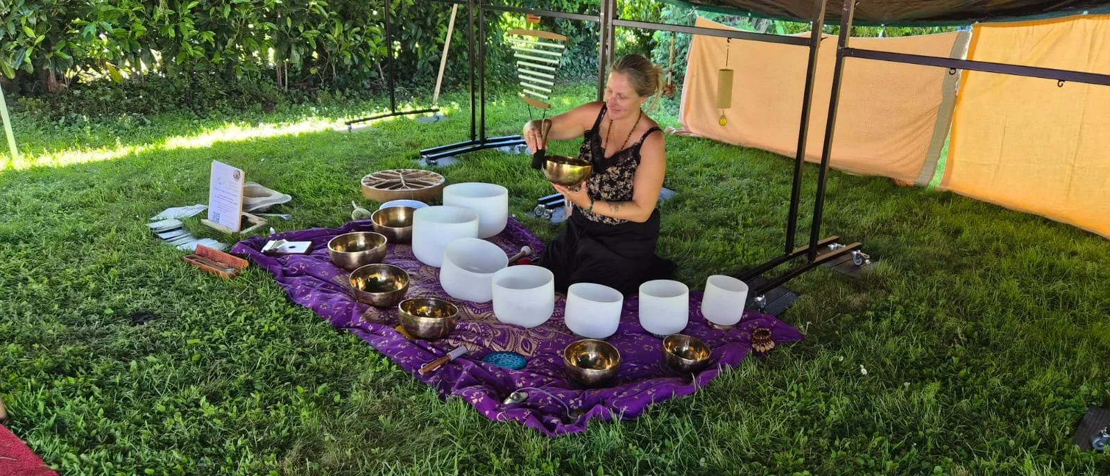 Femme jouant des bols tibétains dans un espace extérieur avec tapis violet, entourée de verdure.