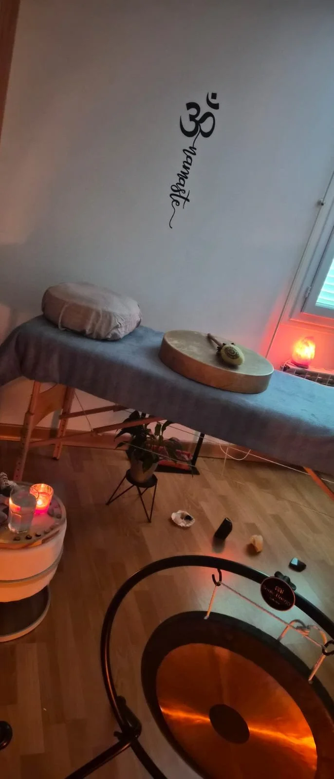 Une salle de massage avec une table de massage recouverte d'un drap gris, un coussin, une pierre polie et un rouleau de massage dessus. Il y a une petite plante en pot, une lampe en forme de cristal orange, et quelques objets sur le sol, dont des pierres. La décoration comprend un mur avec le symbole Om et le mot 'Namaste' écrit verticalement. Il y a aussi un gong en cuivre dans la pièce.