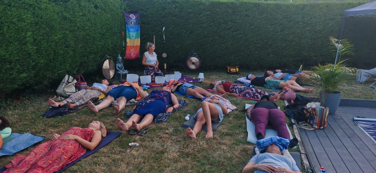 Groupe de personnes pratiquant la méditation ou la relaxation sur l'herbe dans un jardin, avec une femme assise au centre, entourée de gongs, gongas et autres instruments, sous un parasol.
