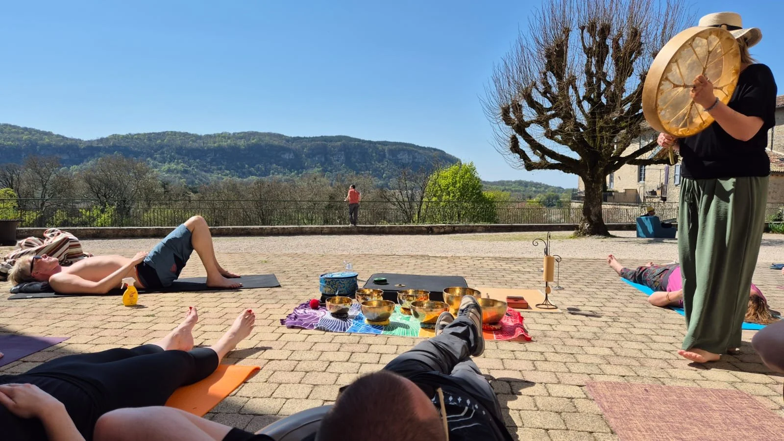 Groupe de personnes pratiquant le yoga ou la méditation en plein air sur une terrasse ensoleillée, avec des montagnes en arrière-plan, une personne jouant d'un instrument, et un arbre nue en toile de fond.