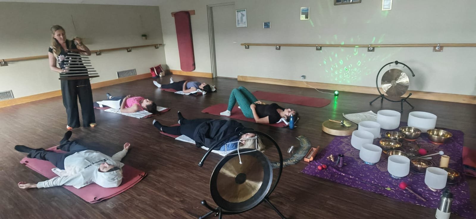 Groupe de personnes faisant une séance de relaxation ou de méditation dans une salle avec des instruments de musique comme des bols tibétains et des gongs, avec des personnes allongées sur des tapis de yoga.