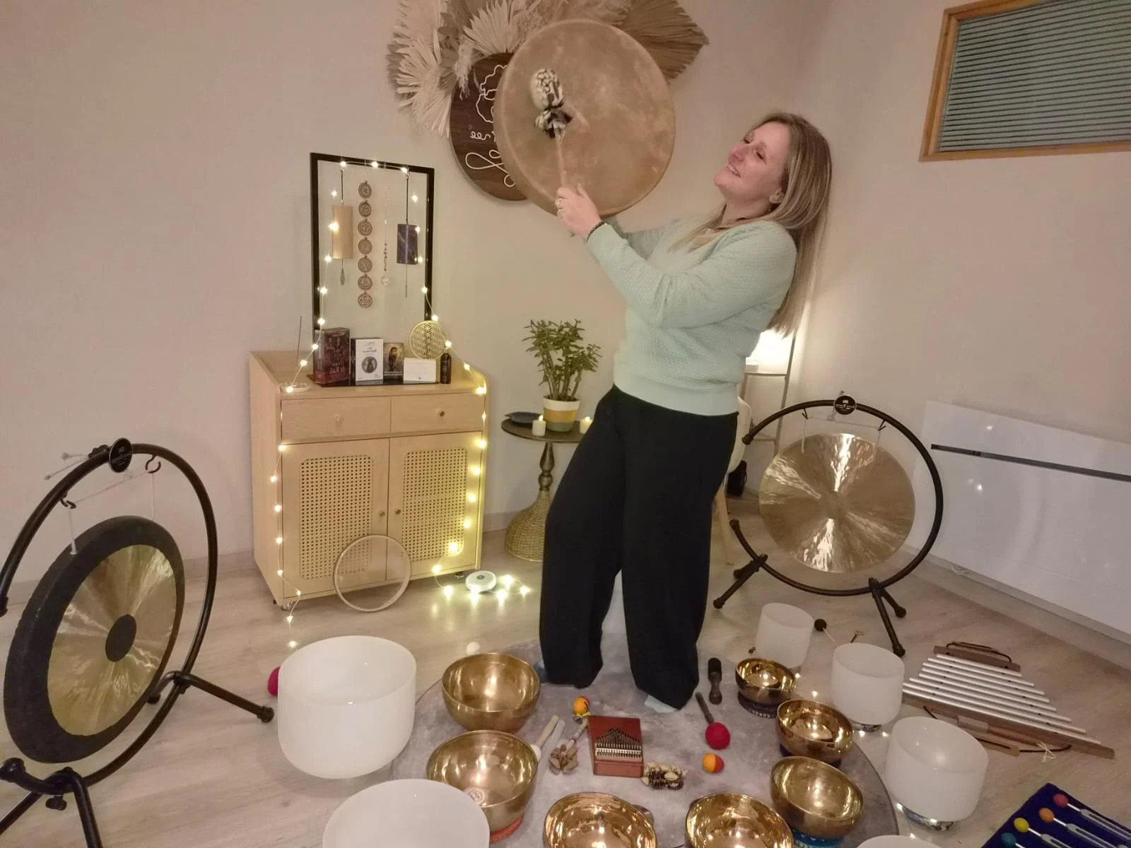 Une femme joue des instruments de musique et des harmonies de sons dans une pièce décorée chaleureusement, avec des bols chantants, sons, guitares et autres objets musicaux décoratifs.