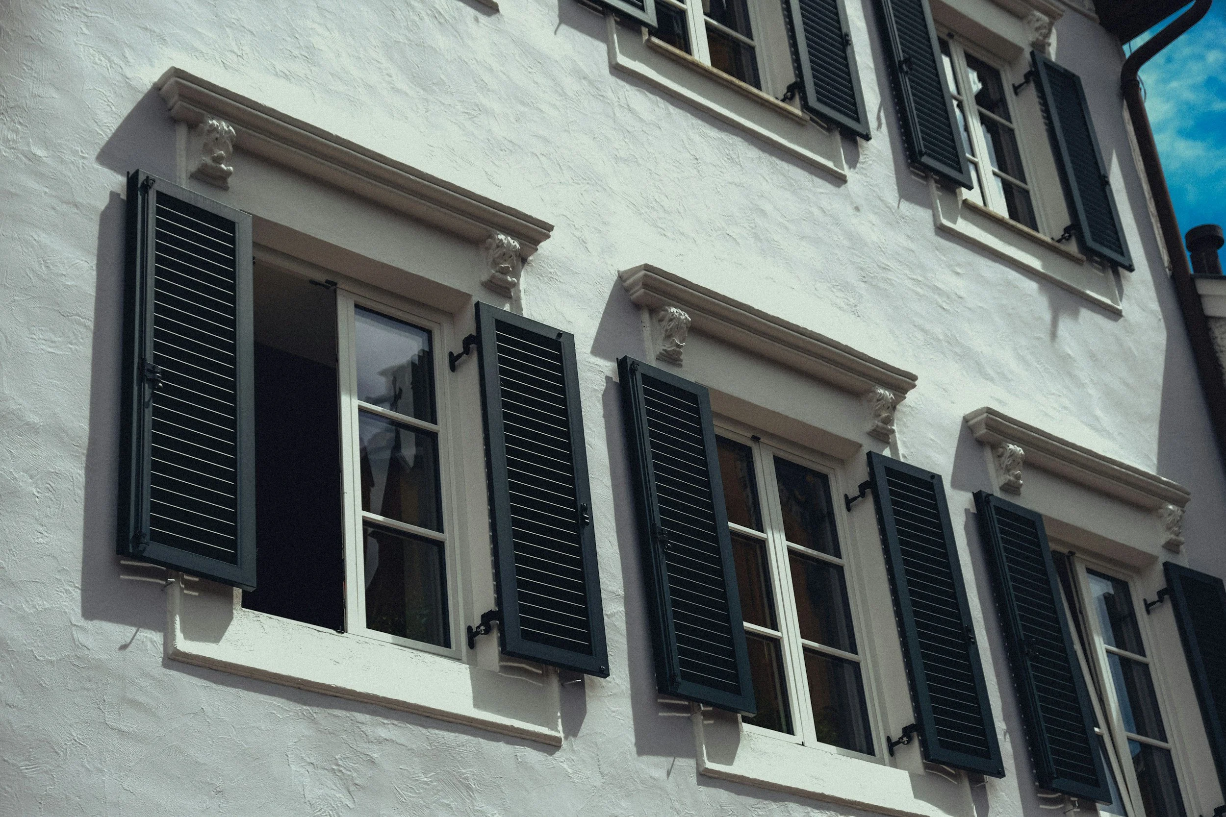 White building facade with multiple windows, each having black shutters, decorative white brackets, and a clear sky in the background.