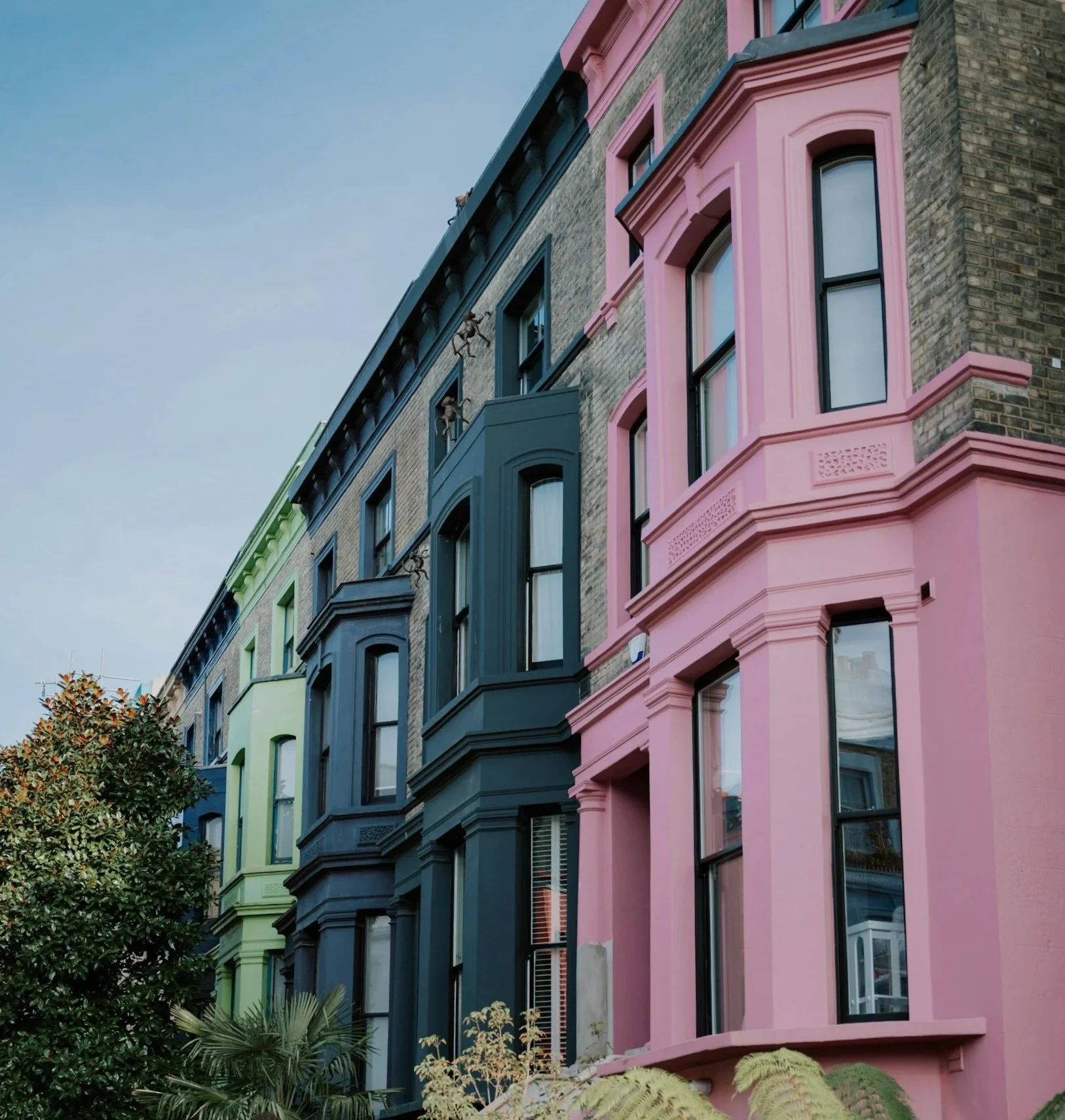 Colorful row of brownstone townhouses with painted facades in pastel colors including pink, blue, green, and black, with bay windows and plants in front.