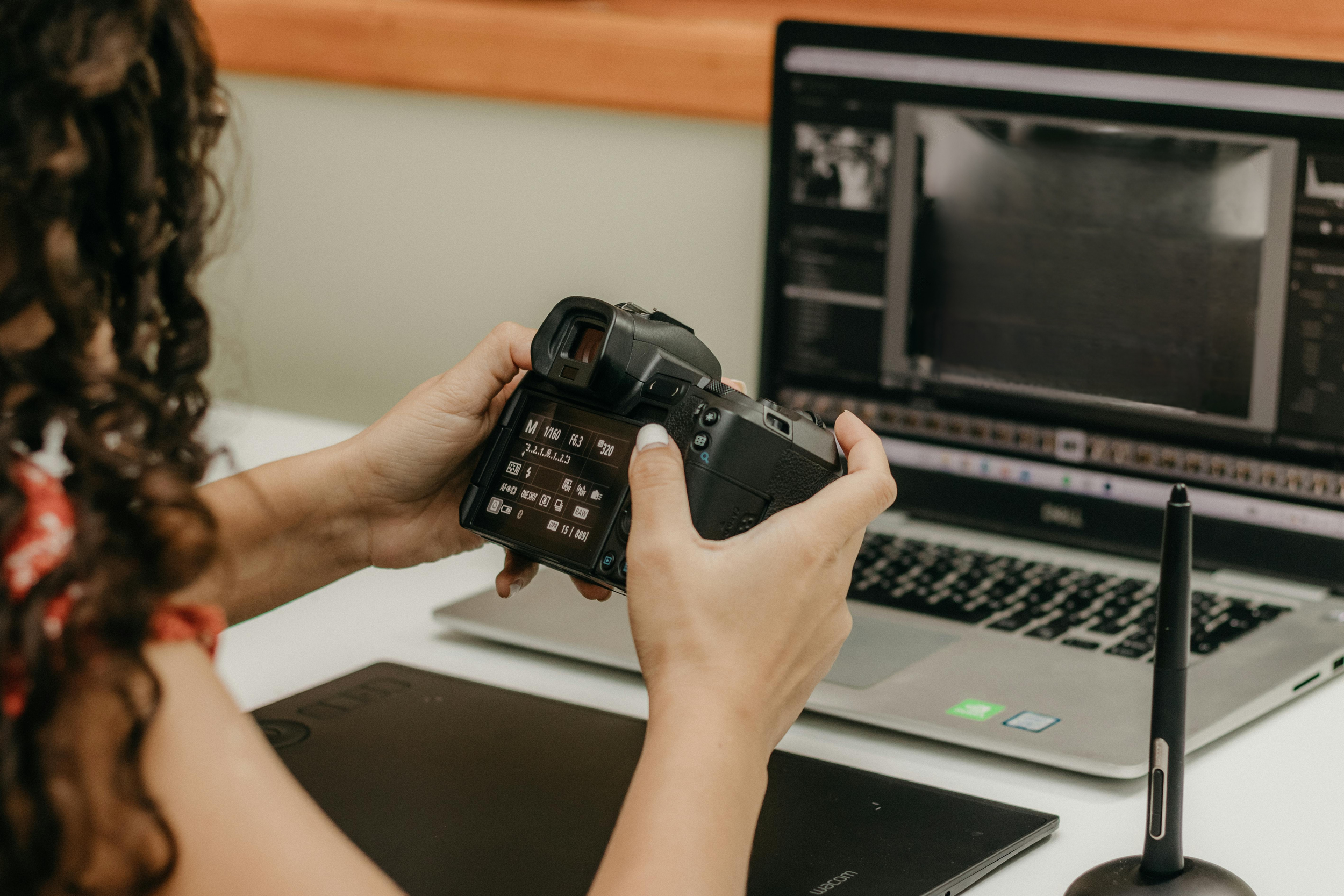Person holding a digital camera in front of a laptop with an editing software open on the screen.