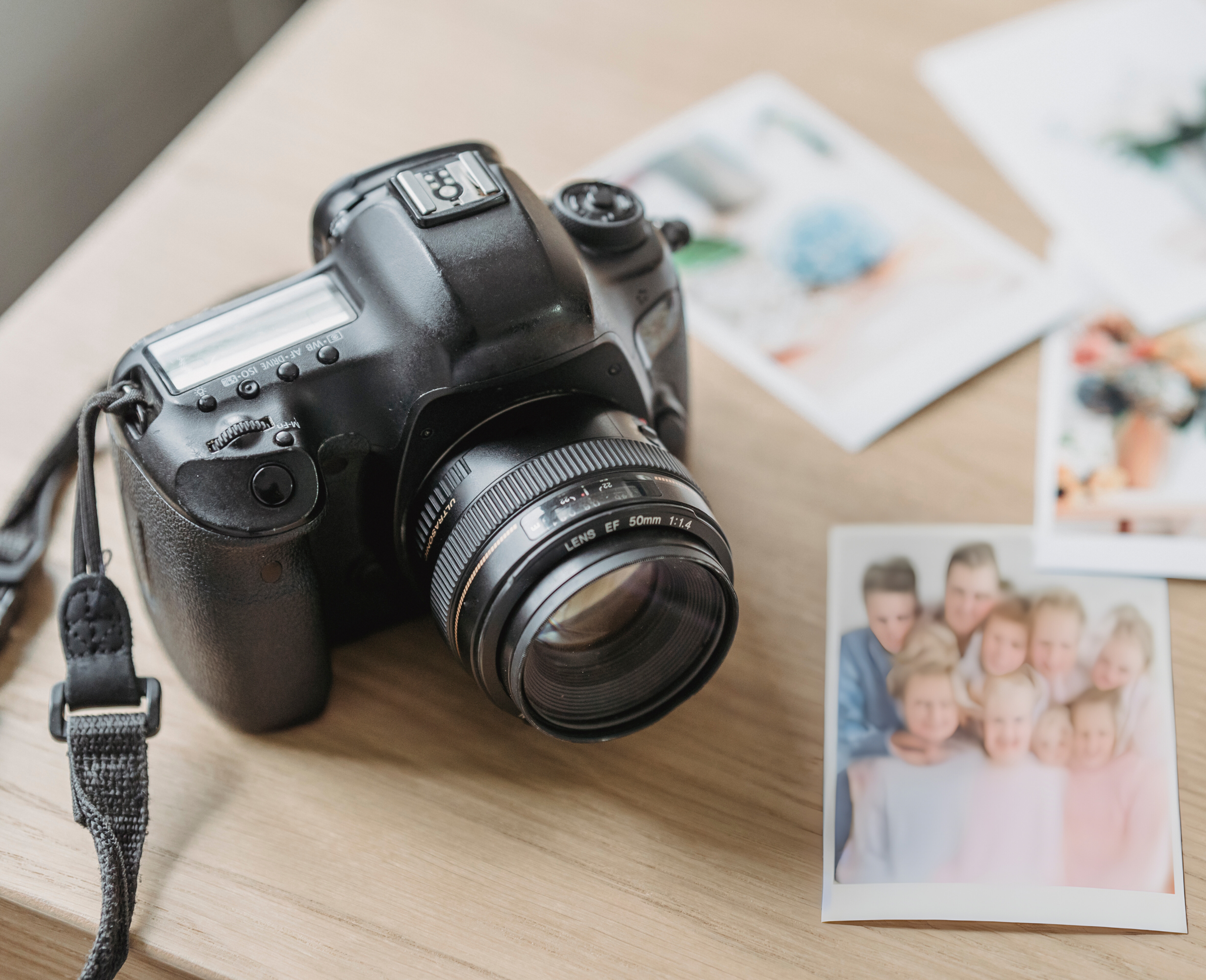 A black digital camera with a lens positioned on a wooden surface surrounded by printed photographs, including family portraits.
