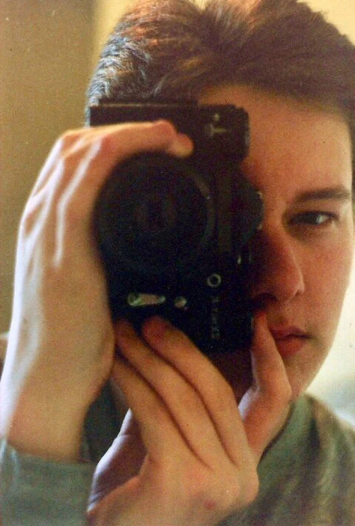 A young person with short dark hair taking a mirror selfie with a black camera, partially covering their face, with one eye open and the other closed or squinting.