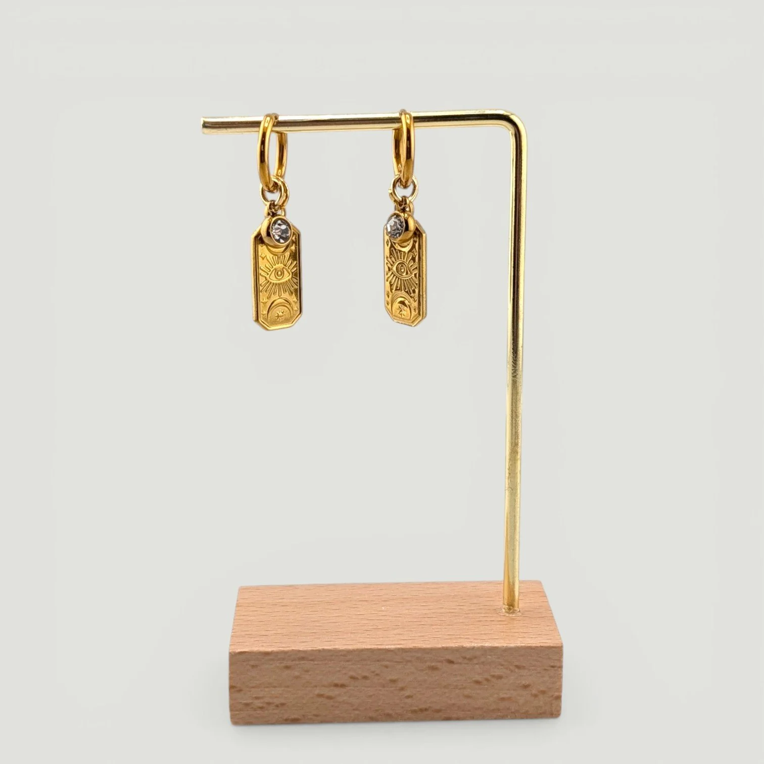 Gold evil eye stainless steel huggie hoop earrings with rhinestone and medallion charms hanging on display stand