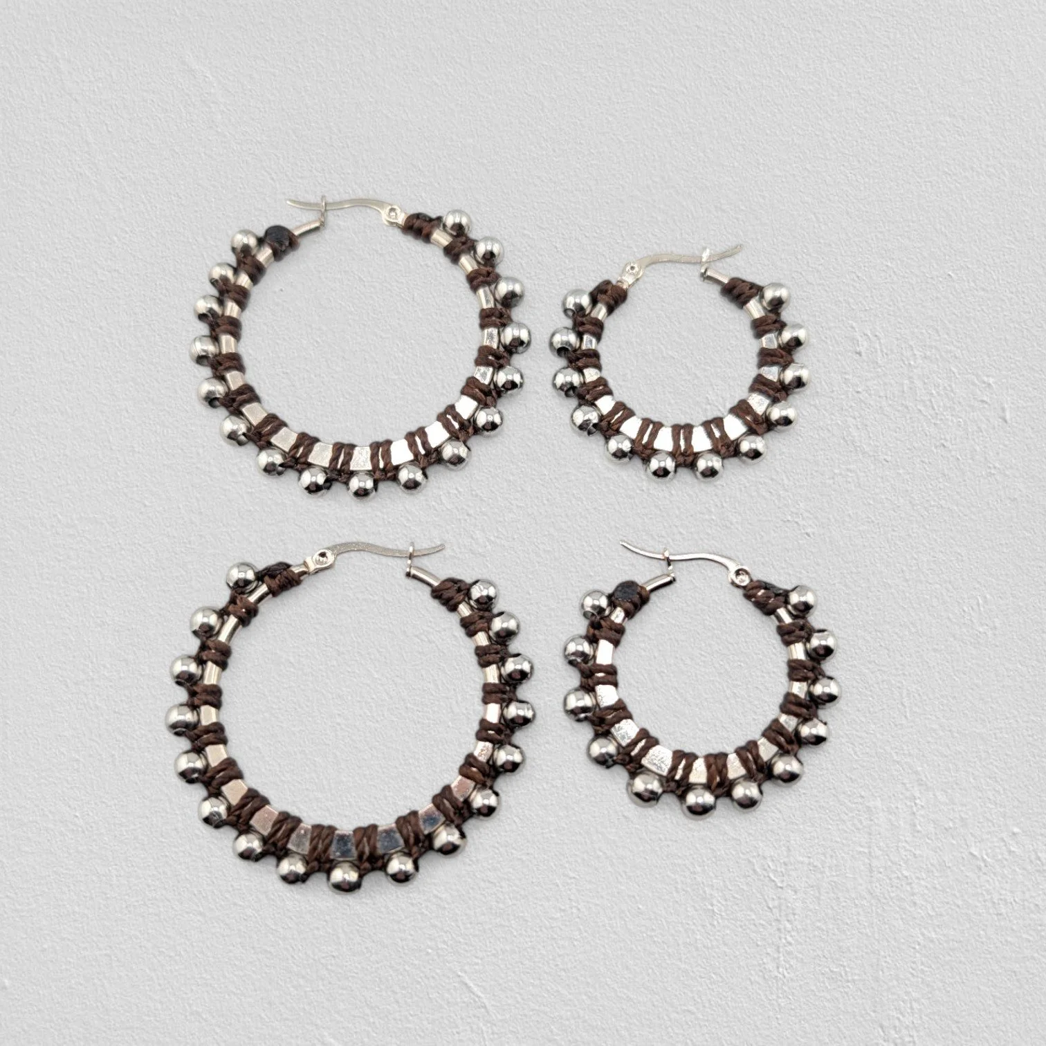 Dark brown beaded macramé stainless steel hoop earrings in silver, 30 mm and 40 mm sizes side by side, flat lay on a neutral background