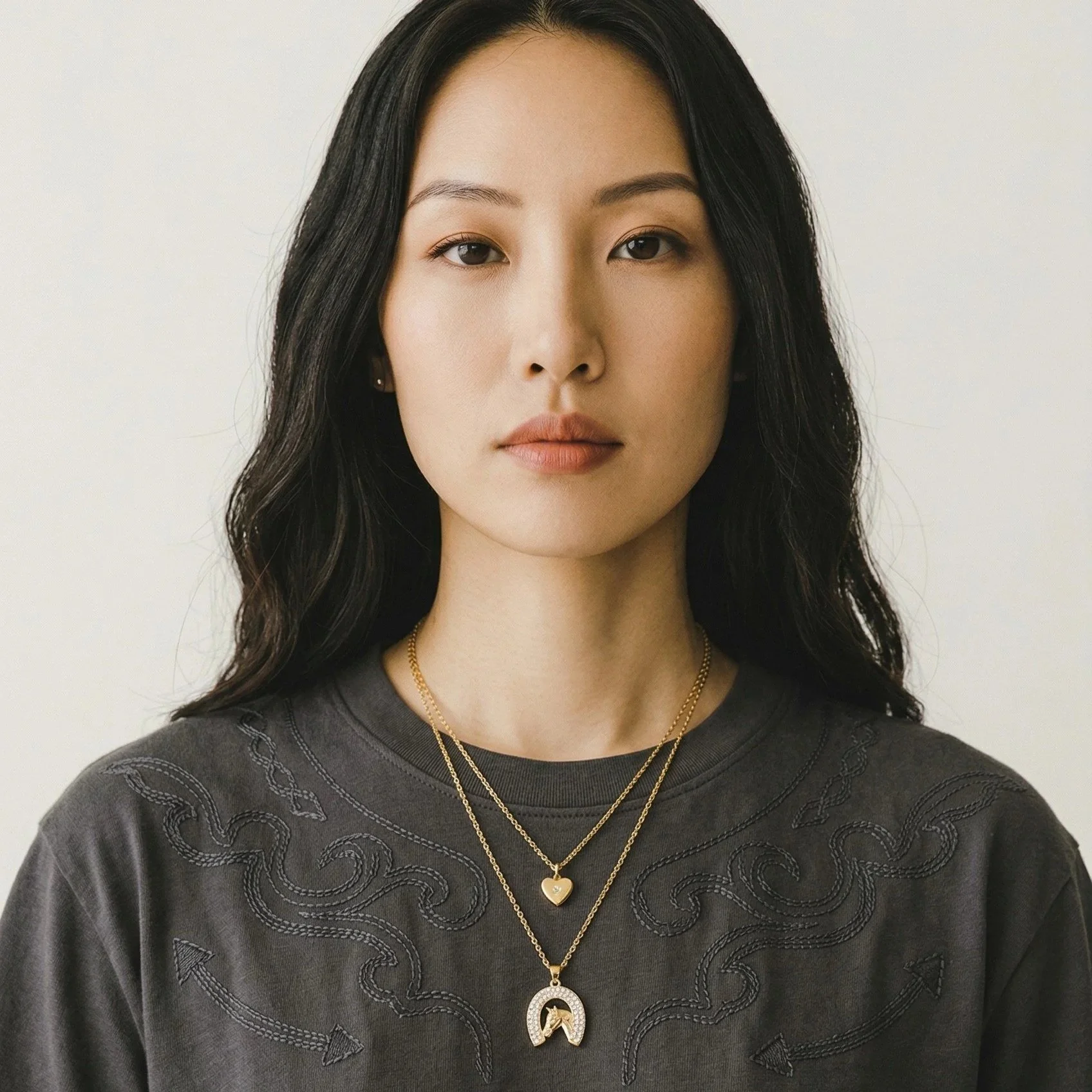 dark haired model wearing rhinestone horseshoe horse gold necklace layered with starburst heart necklace and a charcoal t-shirt