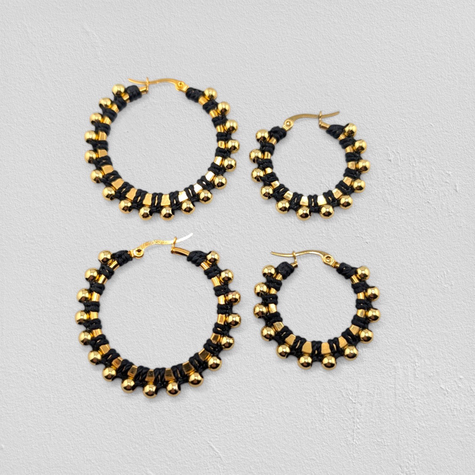 Black beaded macramé stainless steel hoop earrings in gold, 30 mm and 40 mm sizes side by side, flat lay on a neutral background