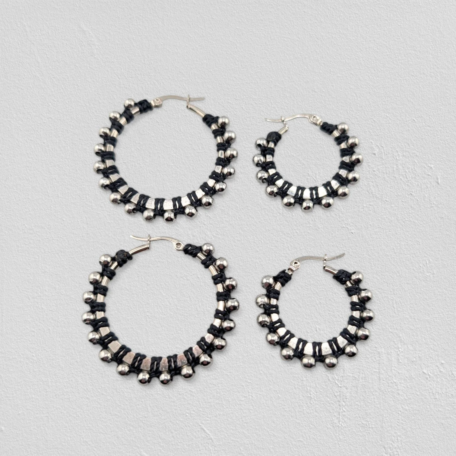 Black beaded macramé stainless steel hoop earrings in silver, 30 mm and 40 mm sizes side by side, flat lay on a neutral background
