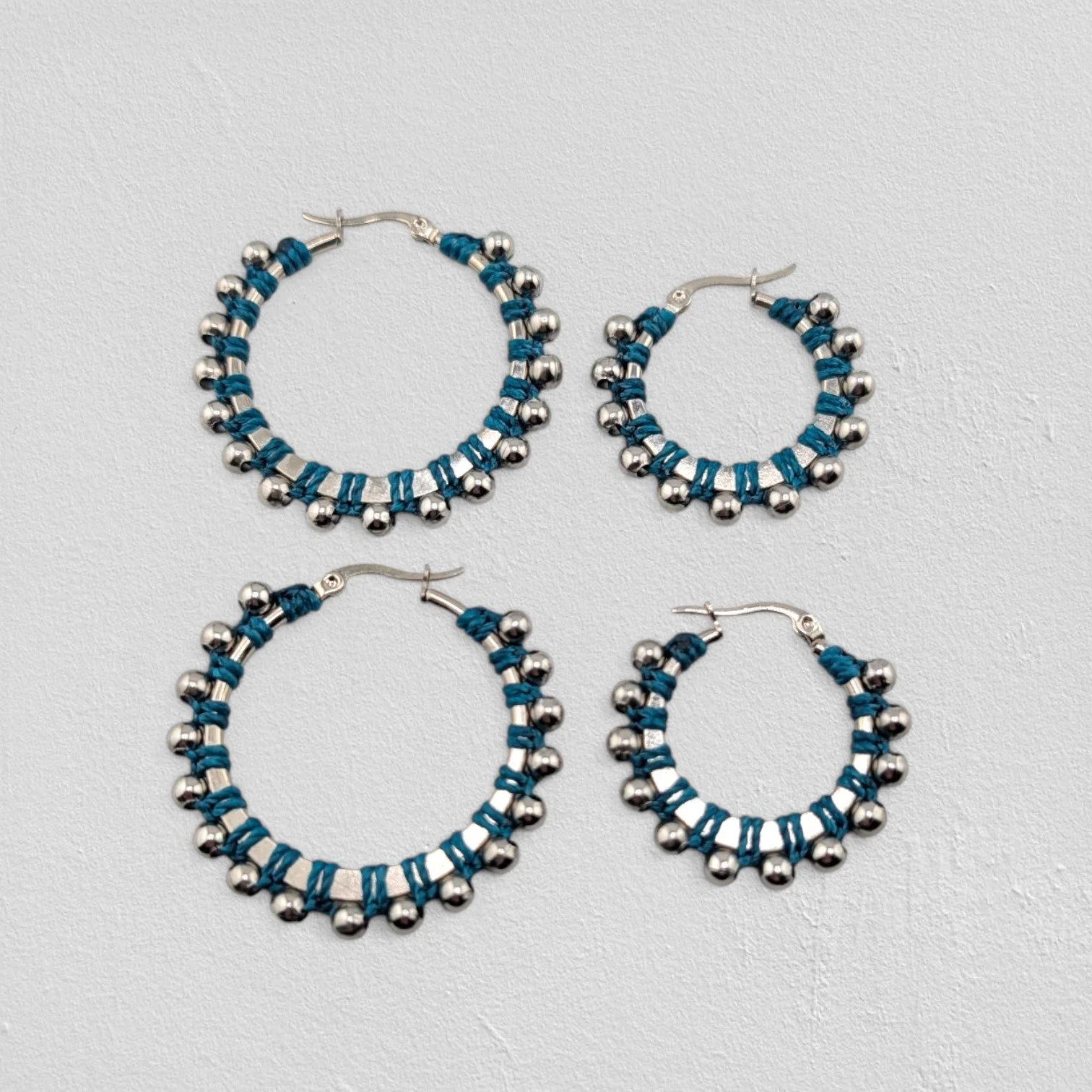 Teal beaded macramé stainless steel hoop earrings in silver, 30 mm and 40 mm sizes side by side, flat lay on a neutral background