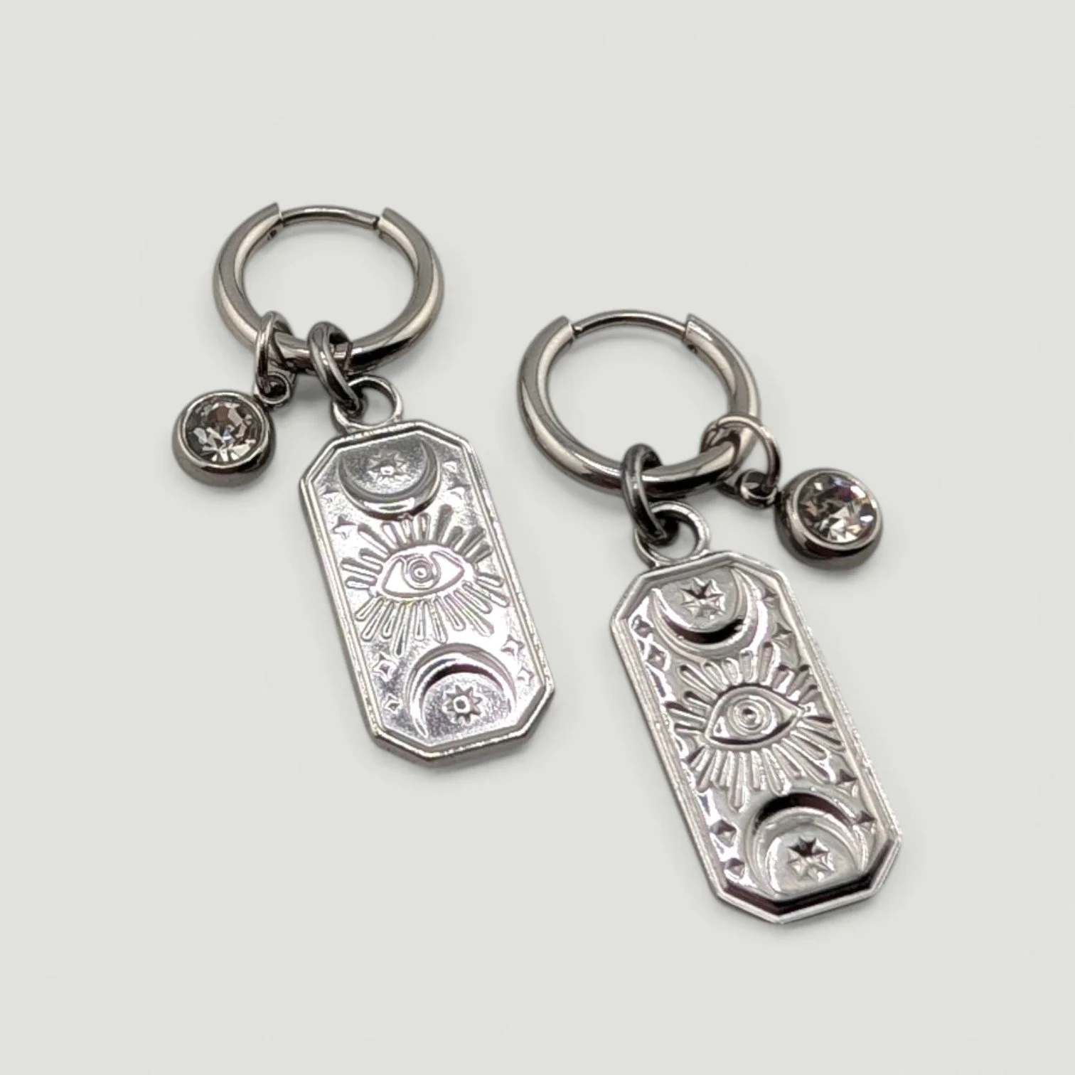 Silver evil eye stainless steel huggie hoop earrings with rhinestone and medallion charms flat lay