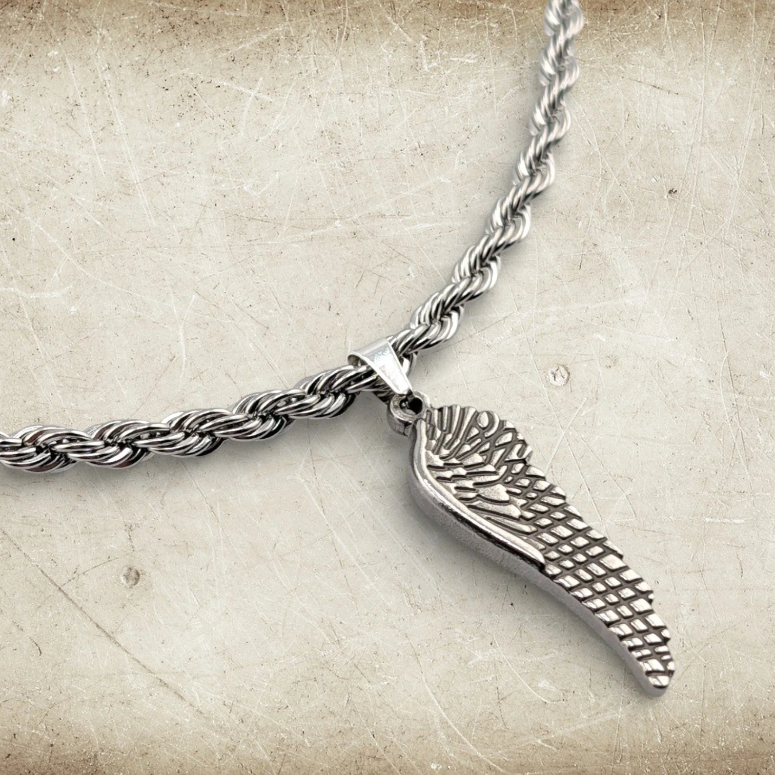 Men's wing stainless steel necklace angled flat lay close-up showing rope chain and pendant on textured background