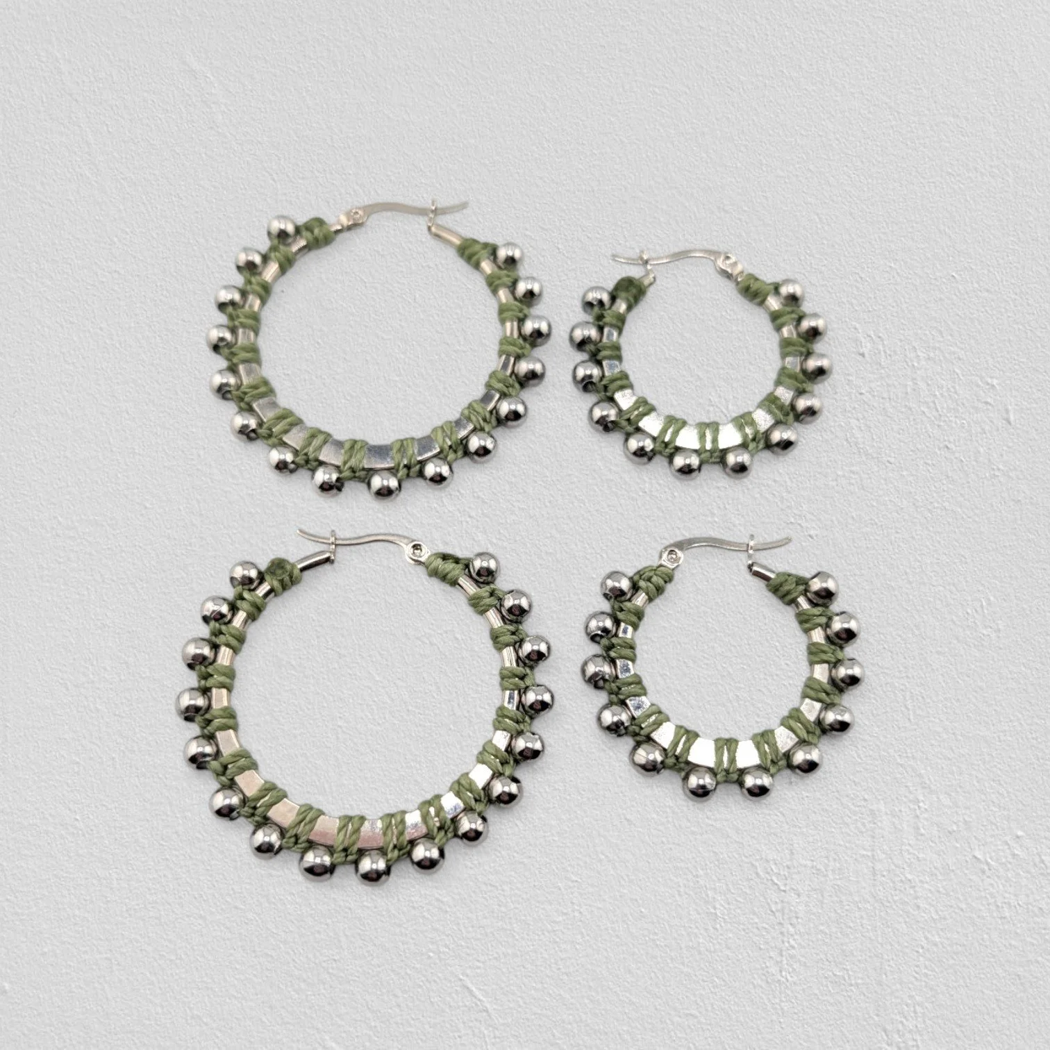 Light moss green beaded macramé stainless steel hoop earrings in silver, 30 mm and 40 mm sizes side by side, flat lay on a neutral background