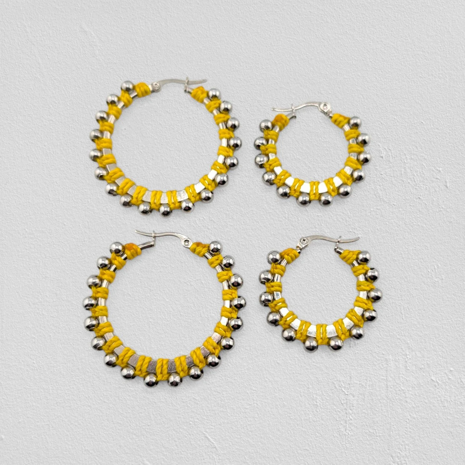 Yellow beaded macramé stainless steel hoop earrings in silver, 30 mm and 40 mm sizes side by side, flat lay on a neutral background