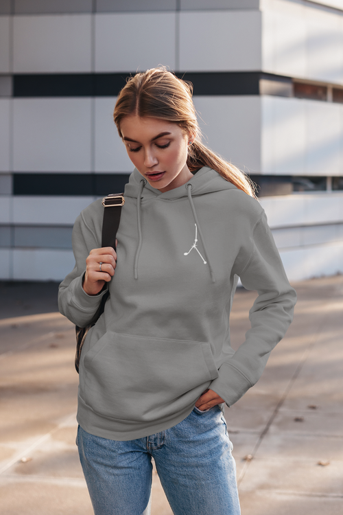 Young woman wearing a gray hoodie and jeans, carrying a backpack, standing outdoors in front of a modern building.