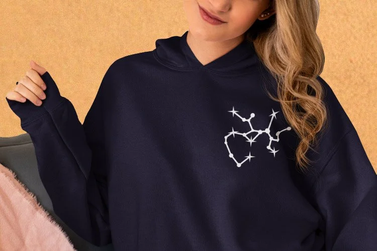 A woman wearing a navy hoodie with a white constellation design on the chest, sitting against a beige background.