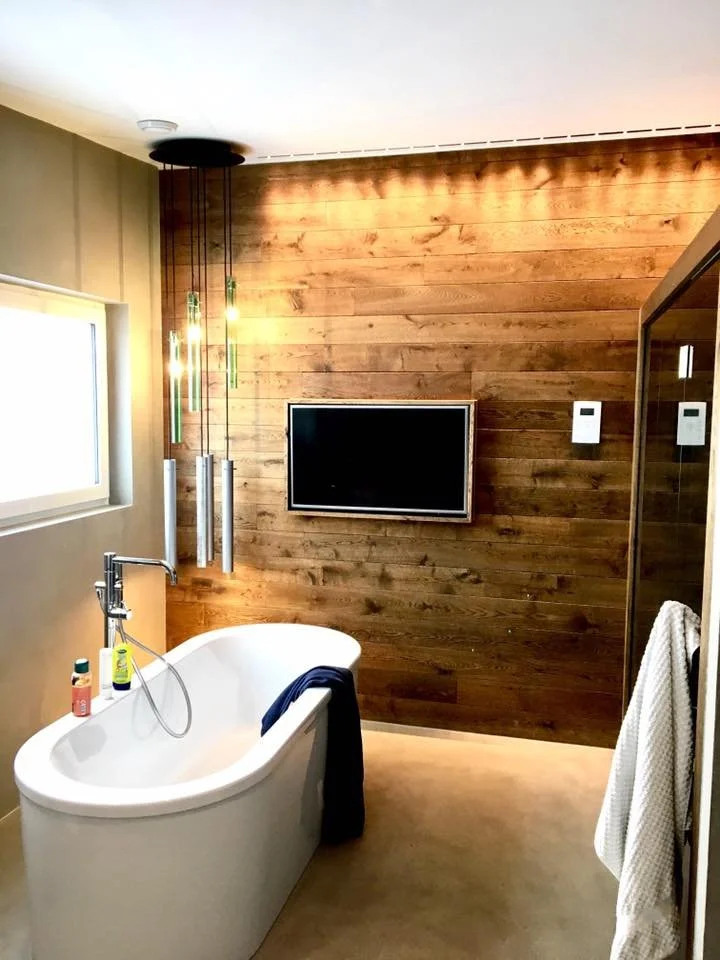  modernes Badezimmer mit freistehender Badewanne, Wand aus Holz, Fernseher an der Wand, Fenster und hängende Beleuchtung