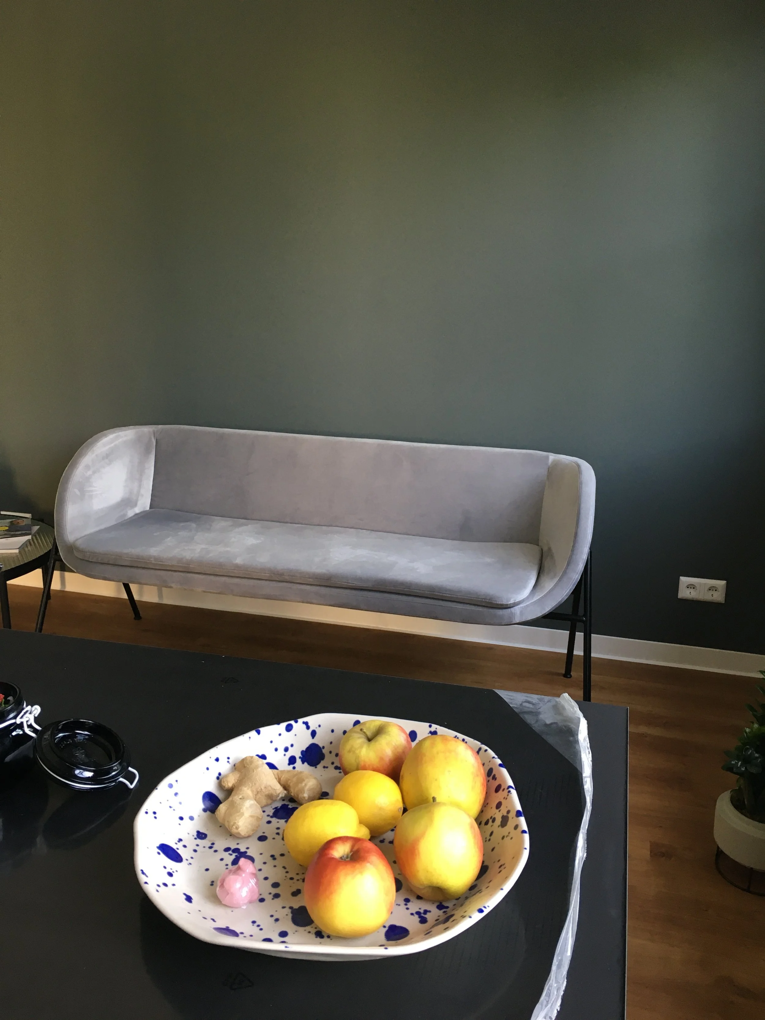 Auf einem schwarzen Tisch liegt eine große weiße Schale mit blauen Sprenkeln, gefüllt mit Äpfeln, Ingwer und einer kleinen rosa Frucht. Im Hintergrund befindet sich ein graues, modernes Sofa vor einer dunkelgrünen Wand.