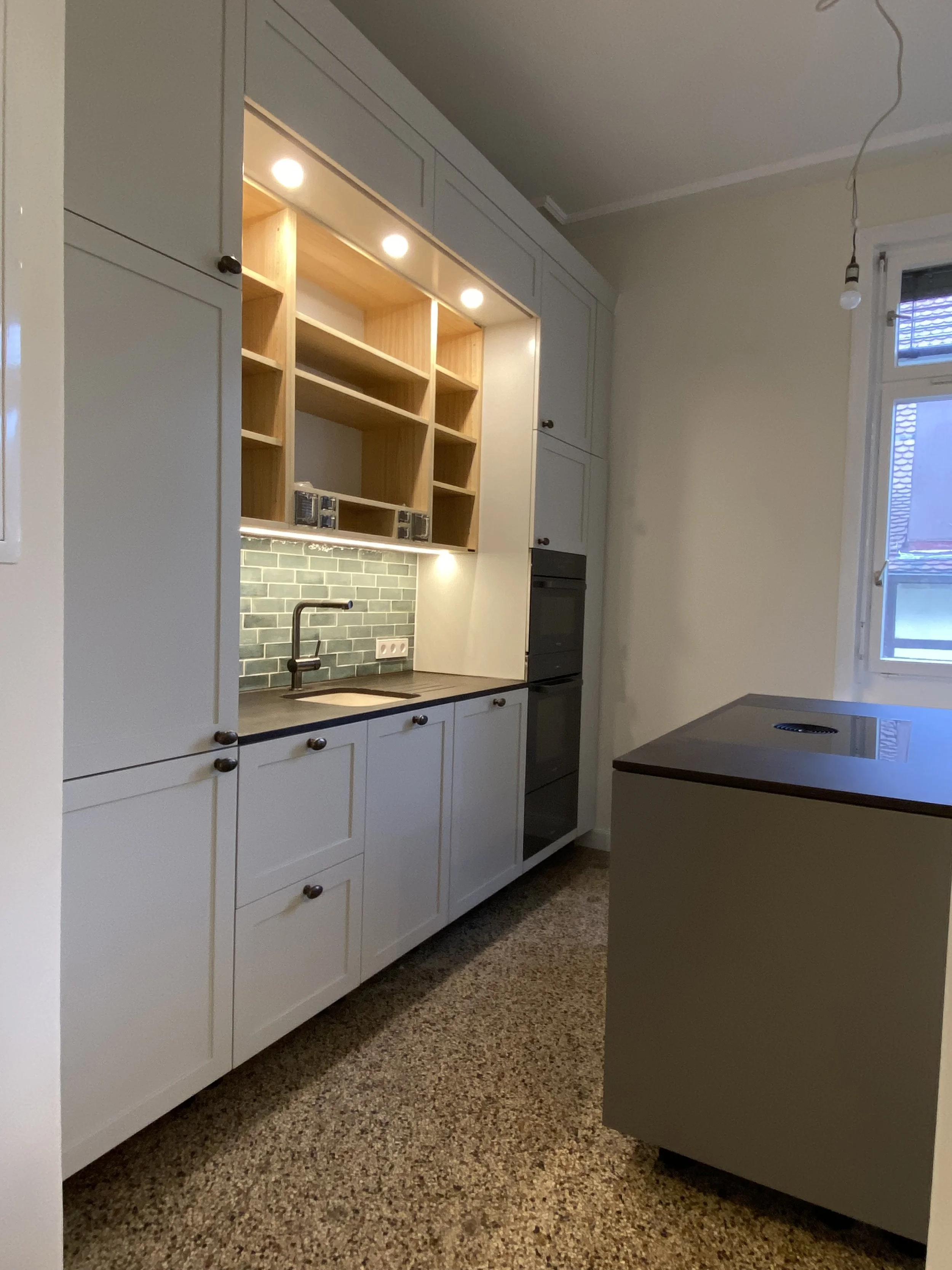 Küchenraum mit weißen Schränken, einem schwarzen Arbeitsplatten, einer grünen Fliesenrückwand, offenen hölzernen Regalen und einem Fenster.