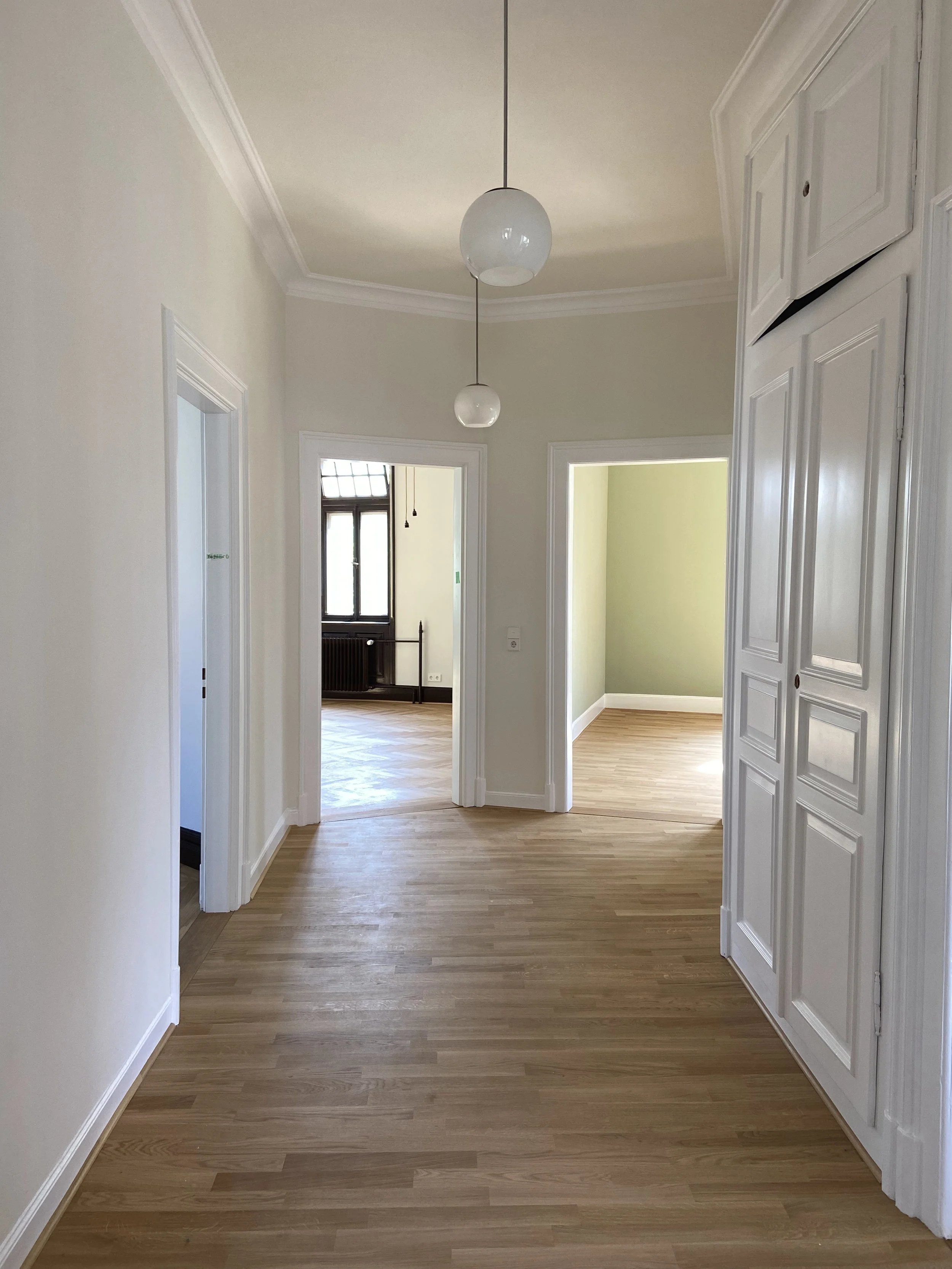 Innenaufnahme eines Flurs in einem Haus mit Holzboden, weißen Wänden und mehreren Türen, zwei große runde Lichtlampen hängen von der Decke