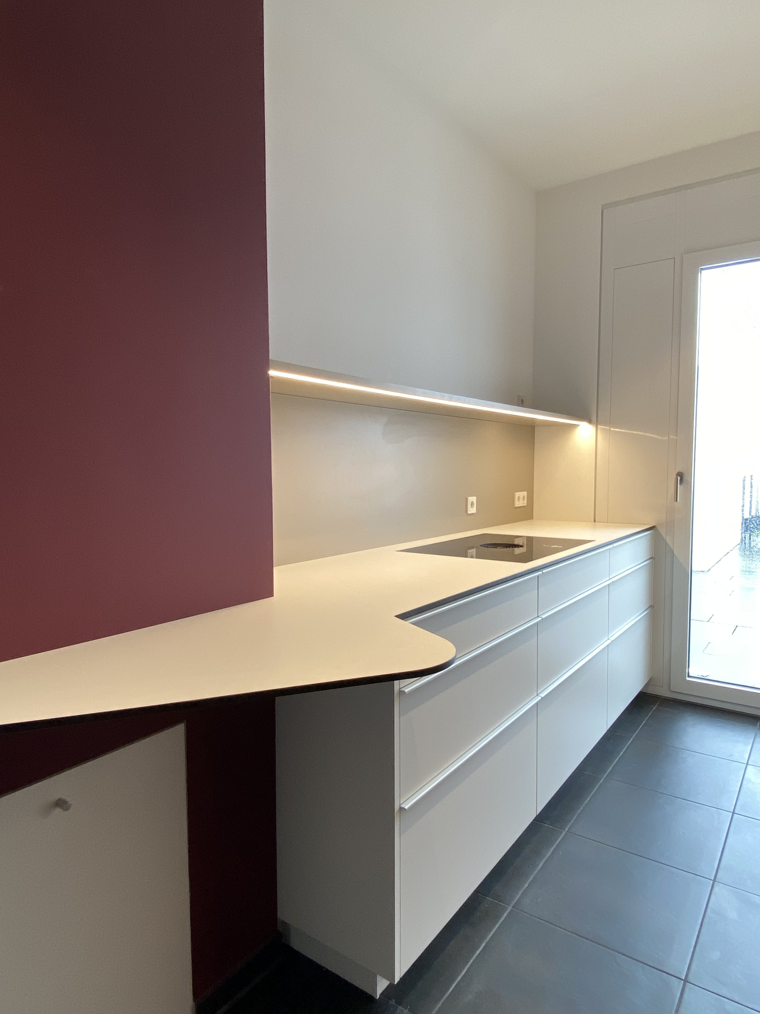 Moderne Küche mit weißen Schränken, grauem Wandschrank, schwarzem Herd, dunklen Fliesenboden und Zugang zu einem Balkon oder Außenbereich.