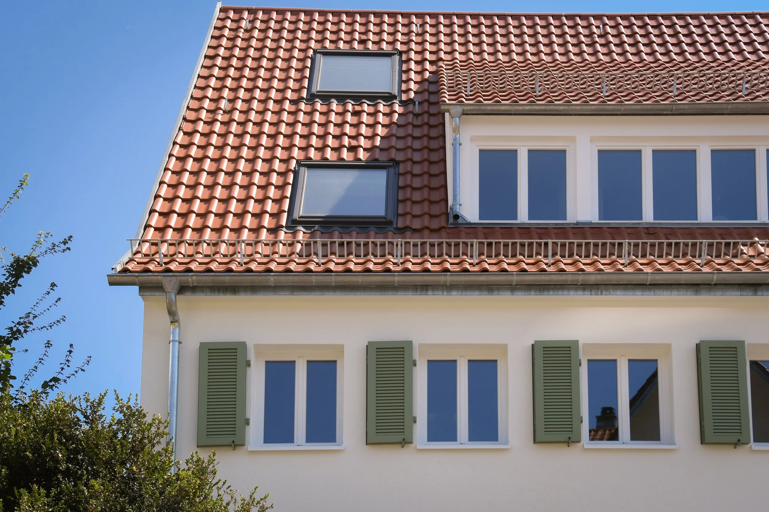 Ansicht eines Mehrfamilienhauses mit roten Schindeln, weißen Fensterrahmen und grünlichen Fensterläden bei blauem Himmel.
