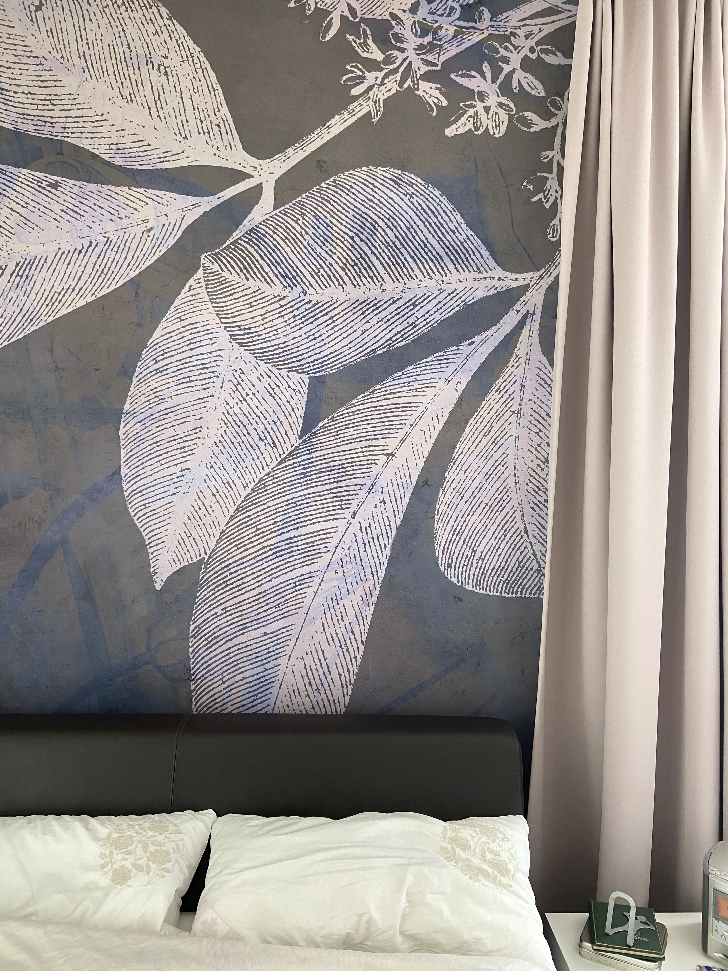 Wanddekoration mit weißen Blättern auf grauem Hintergrund neben beige Vorhängen und einem Bett mit weißen Kissen.