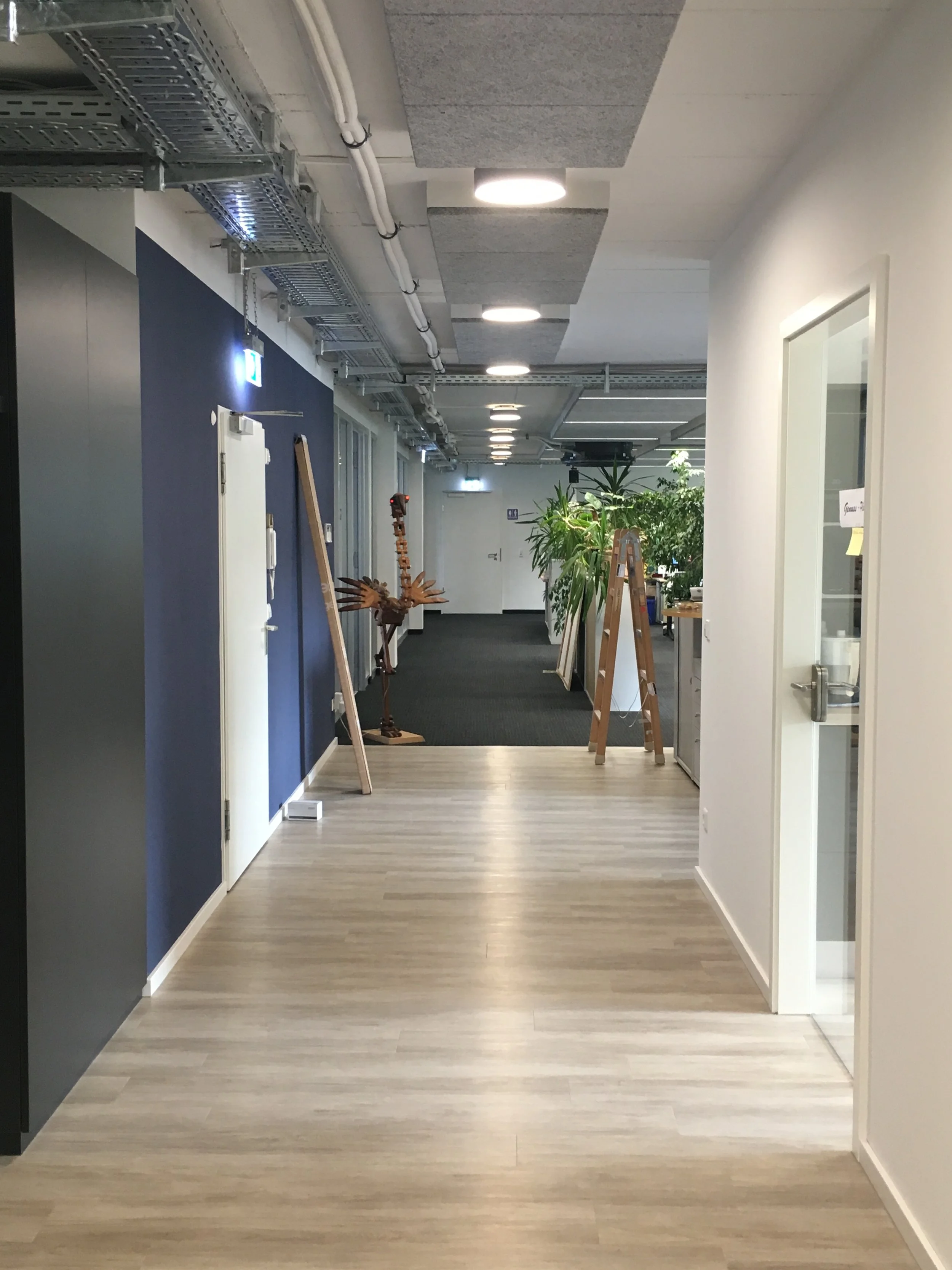 Innenansicht eines Büros mit Flur, Pflanzen, Leitungen an der Decke und einen Stehpult.