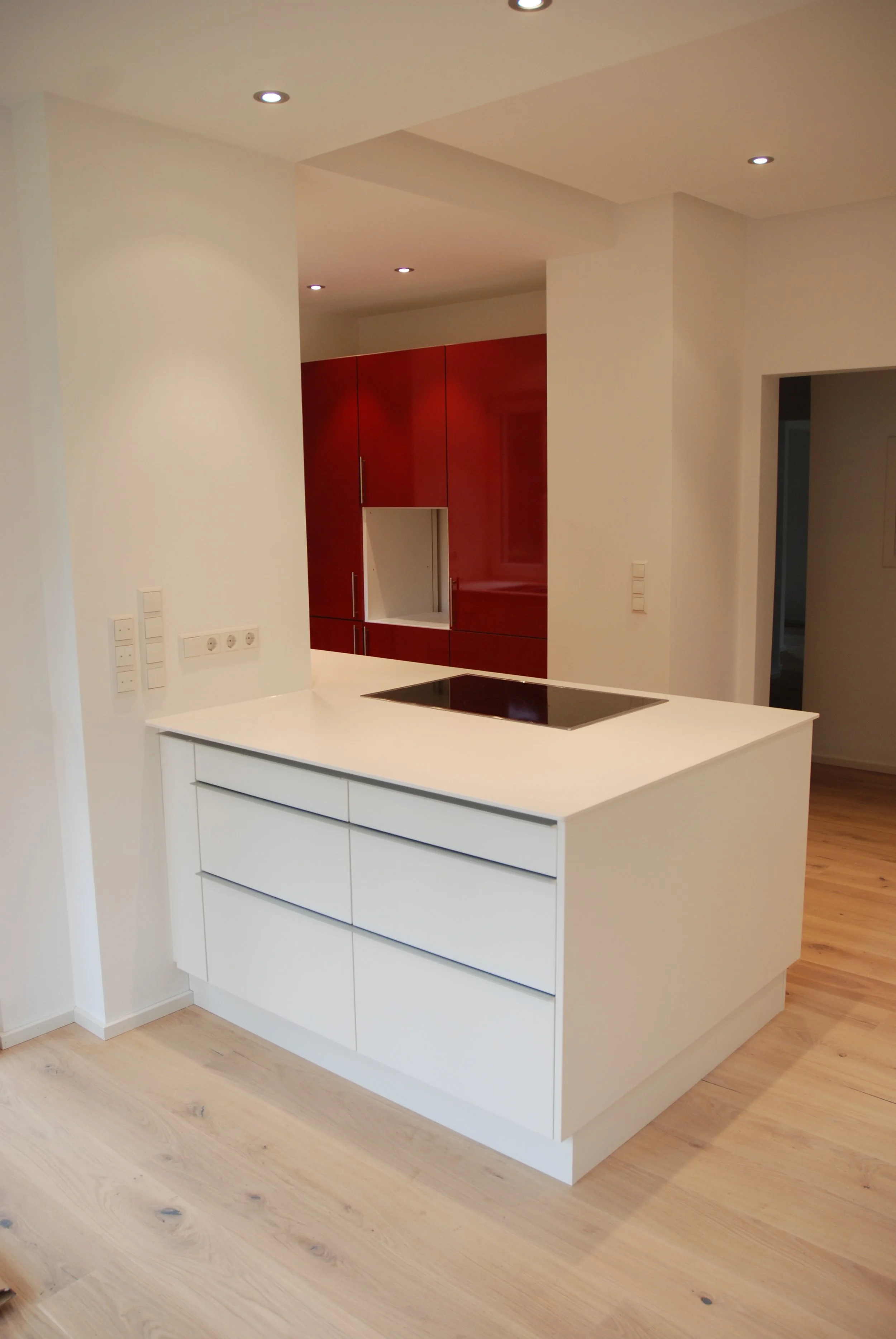 Moderne weiße Kücheninsel mit weißen Schubladen, schwarze Kochplatte und hellem Holzboden in einer Küche mit weißen Wänden und rot glänzenden Küchenschränken im Hintergrund.