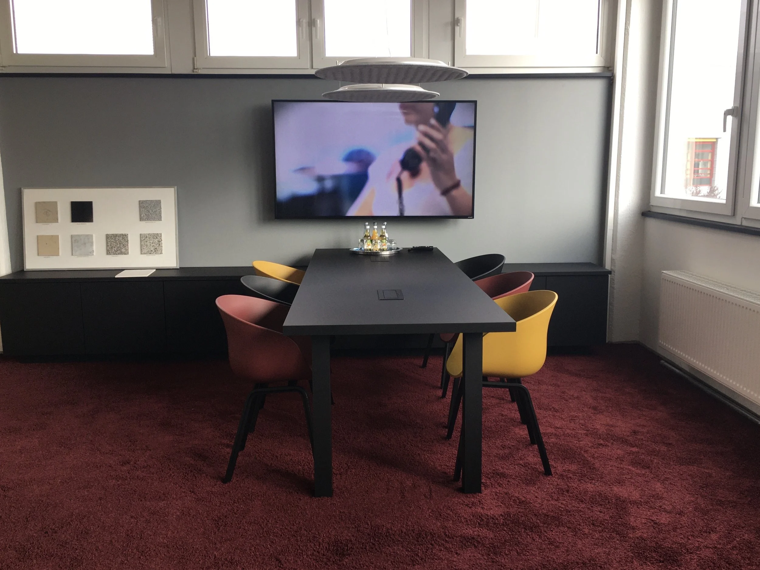 Konferenzraum mit schwarzen Tisch, bunten Stühlen, Fernseher an der Wand, Fenster und rotem Teppich.
