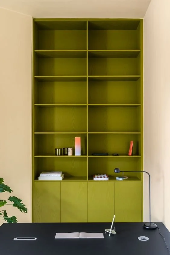 Grüner offene Regalwand an einer weißen Wand, mit einigen Dekorationsgegenständen und Büchern, darunter eine Stehlampe, auf einem schwarzen Tisch davor.