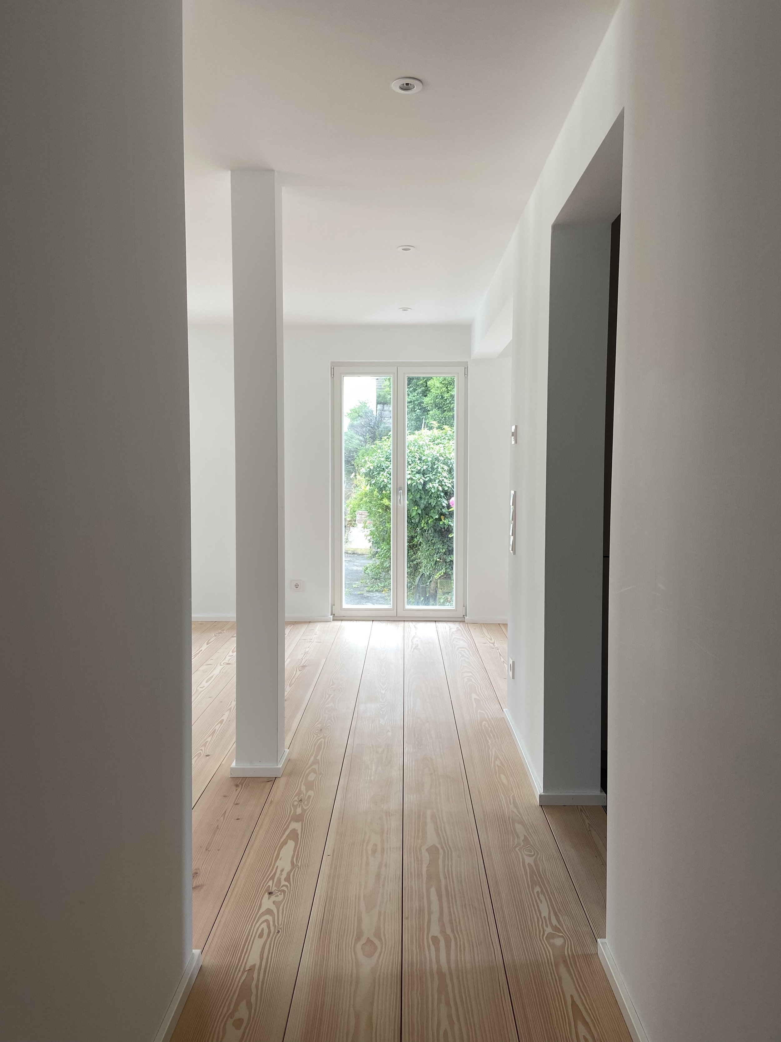 Leeres, helles Zimmer mit weißen Wänden, Holzboden, einem großen Fenster mit Blick auf grüne Bäume, und einer Türöffnung auf der rechten Seite.