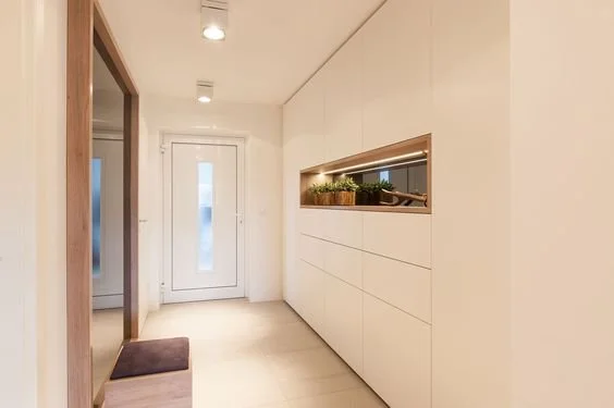 Ein moderner Flur mit weißen Wänden, Holztüren und einem eingebauten Wandregal mit Pflanzen, beleuchtet von Deckenlampen.