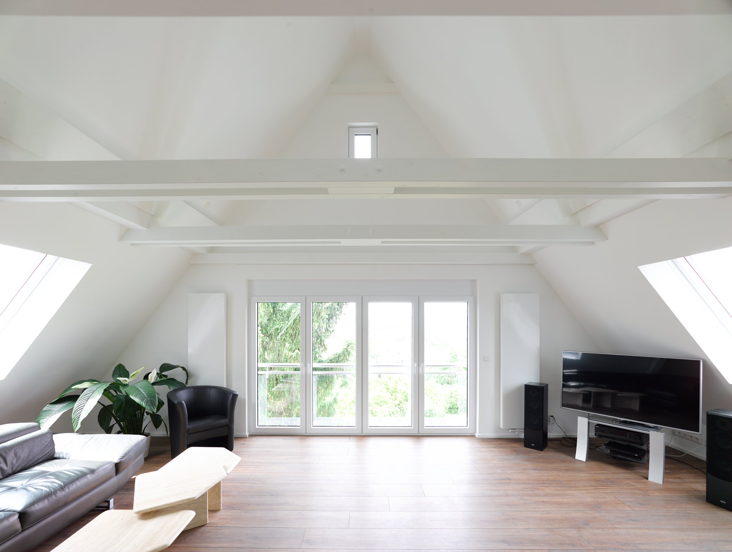 Helles Wohnzimmer mit großen Fenstern, Holzfußboden, schwarzer Sessel, Sofa, großer Flachbildfernseher, Pflanzen und helle Wände.