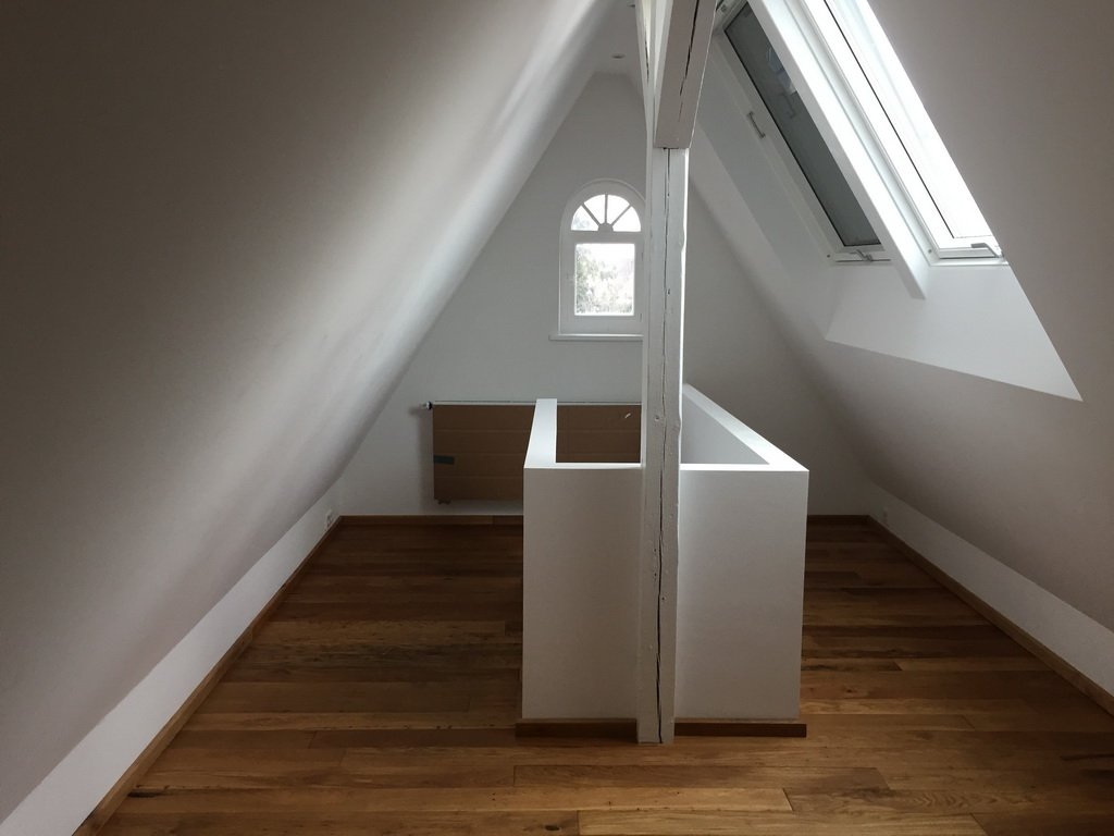 Ein leerer Dachboden mit weißen Wänden, Holzboden und Dachfenstern, in dem eine weiße halbhohe Trennwand steht.