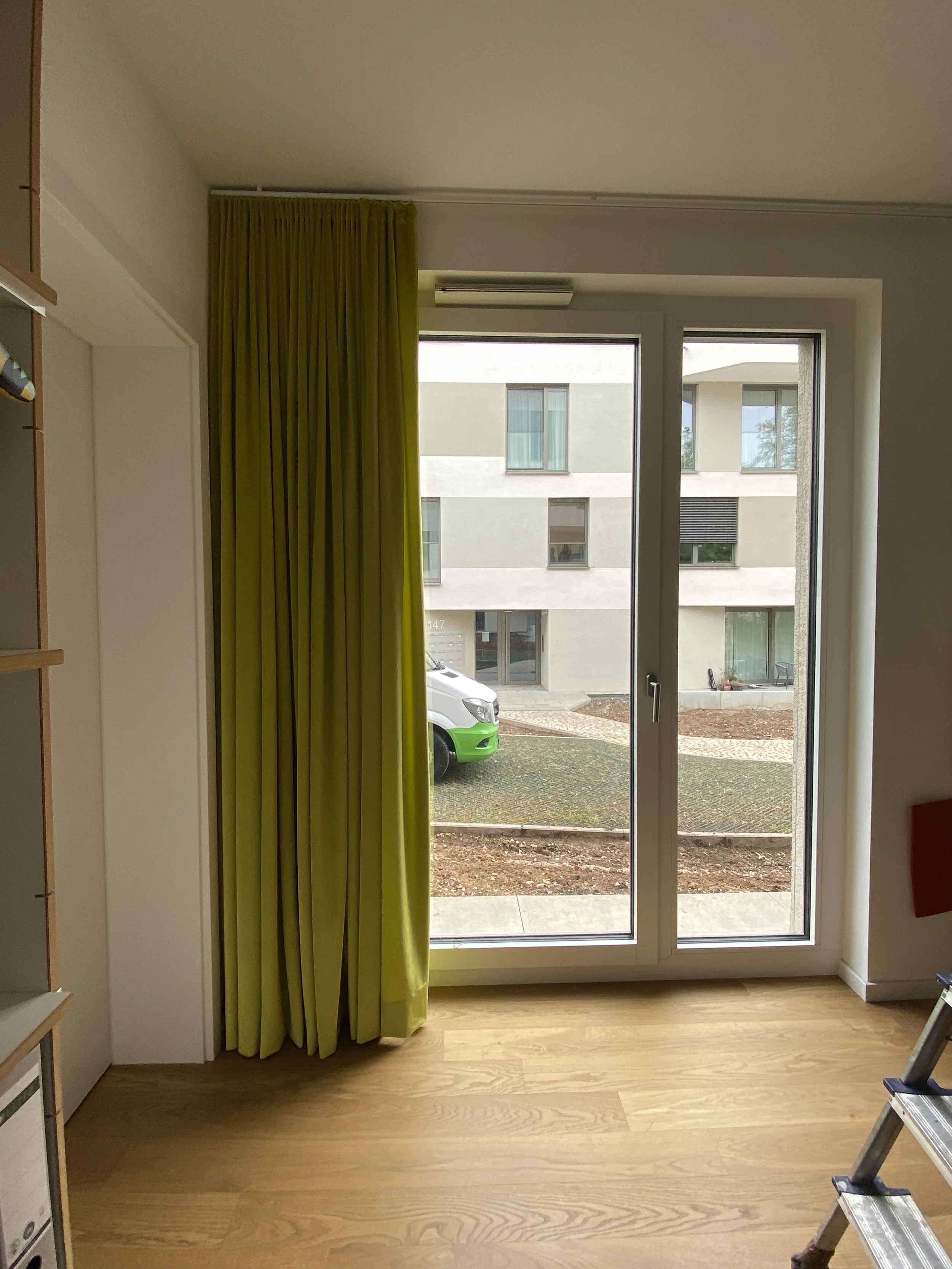 Blick auf ein großes Fenster mit grüner Gardine, Vorhänge sind zur Seite gezogen, mit Blick auf eine Straße und ein modern gestaltetes Mehrfamilienhaus.