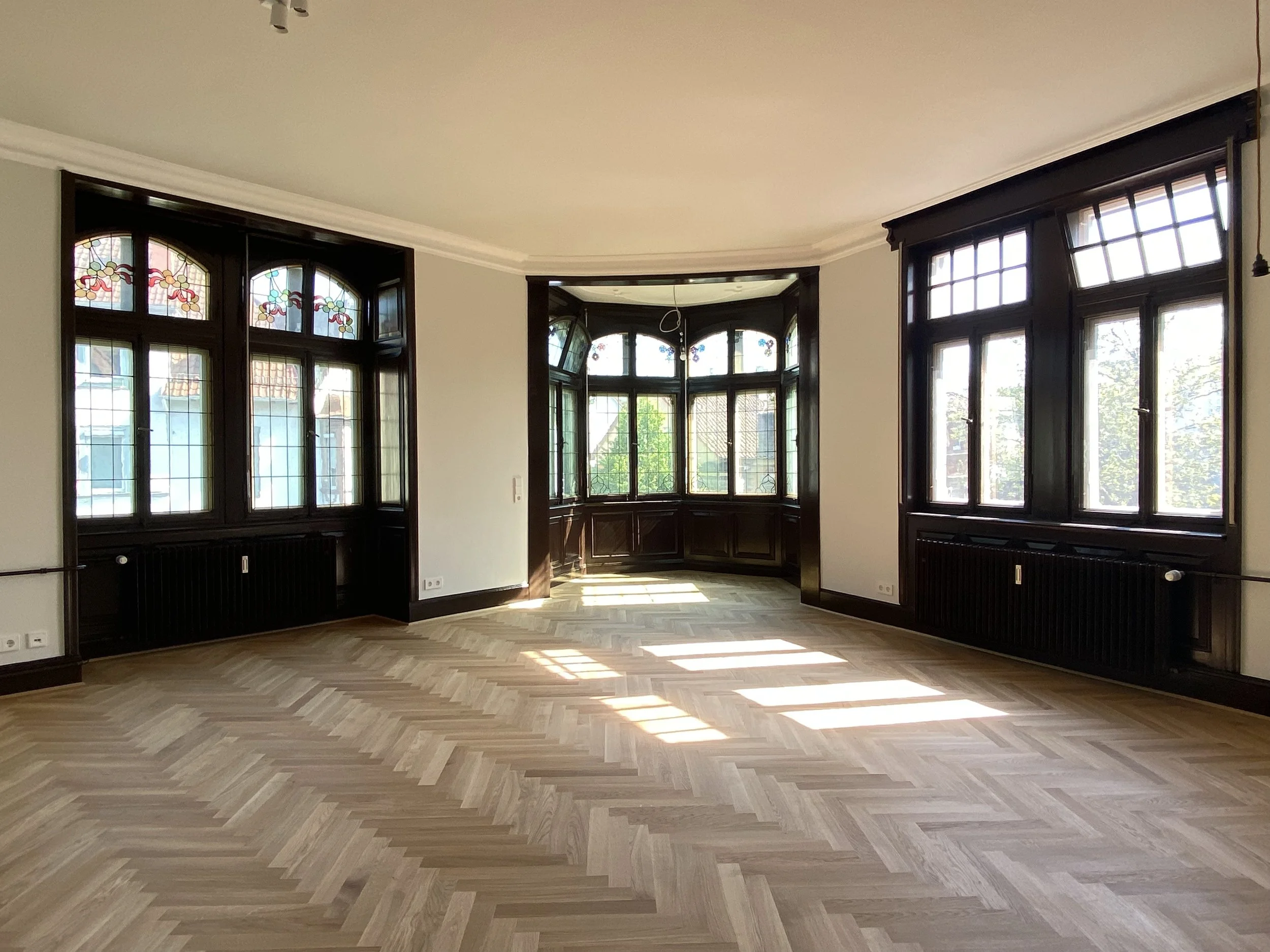 Ein leeres Zimmer mit großen Fenstern, dunklen Holzwänden und Parkettboden, das Sonnenlicht hereinlässt.