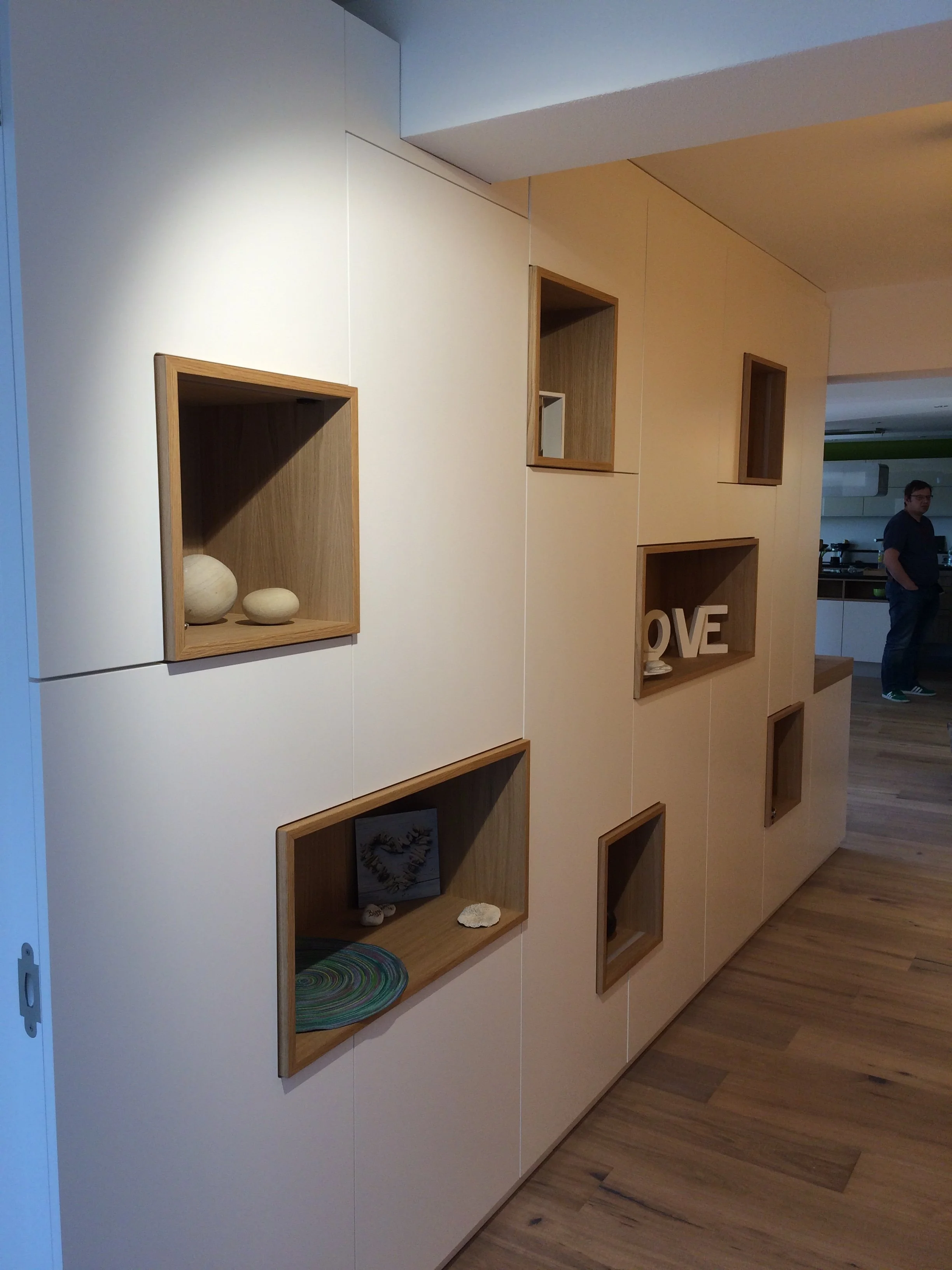 Moderner Raum mit weißen und holzfarbenen Wandregalen, die mit Dekorationsgegenständen und Buchstaben geschmückt sind. Man sieht eine Person im Hintergrund in einer Küche.