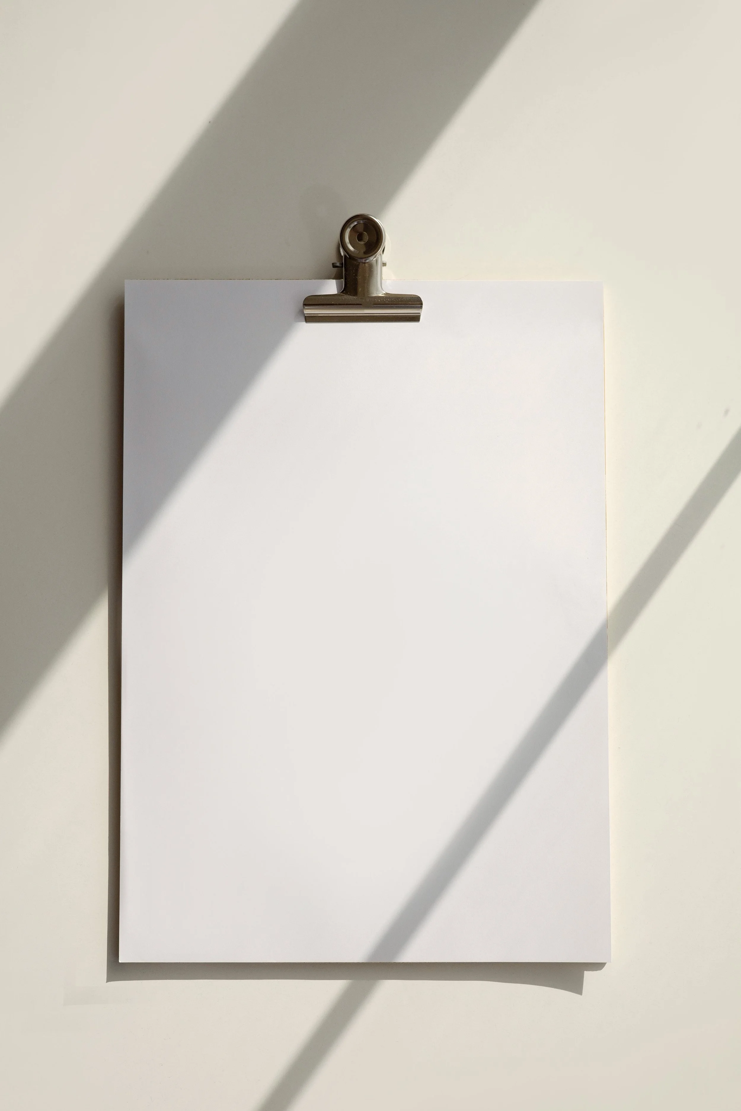A blank white sheet of paper held by a metal clip on a white surface, with shadows cast across the paper.