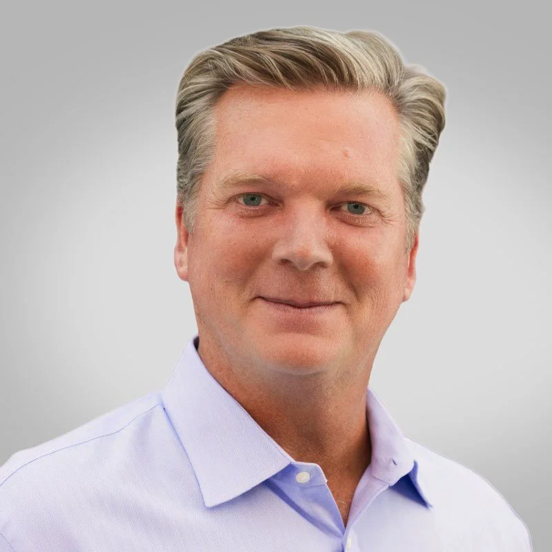 A middle-aged man with light hair and blue eyes smiling wearing a light blue collared shirt against a plain gray background.