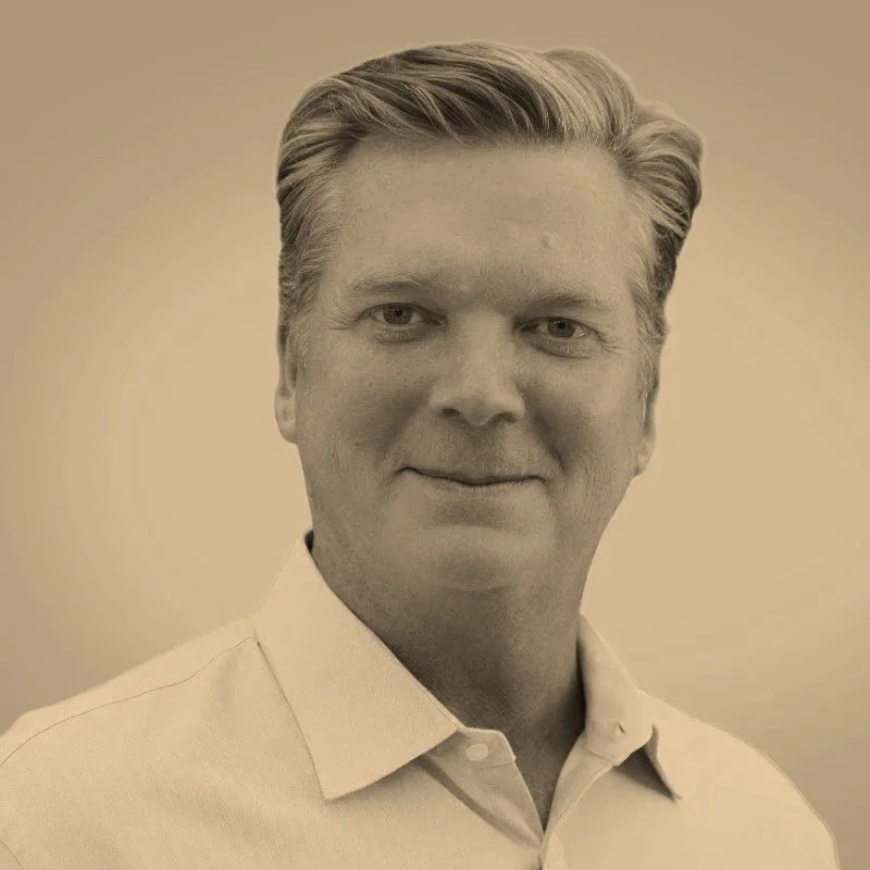 A portrait of a middle-aged man with styled light hair, wearing a white collared shirt, smiling slightly, against a plain beige background.