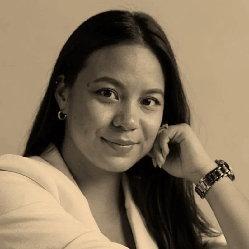 Black and white portrait of a woman with long dark hair, smiling, resting her chin on her hand, wearing a blazer, earrings, and a wristwatch.