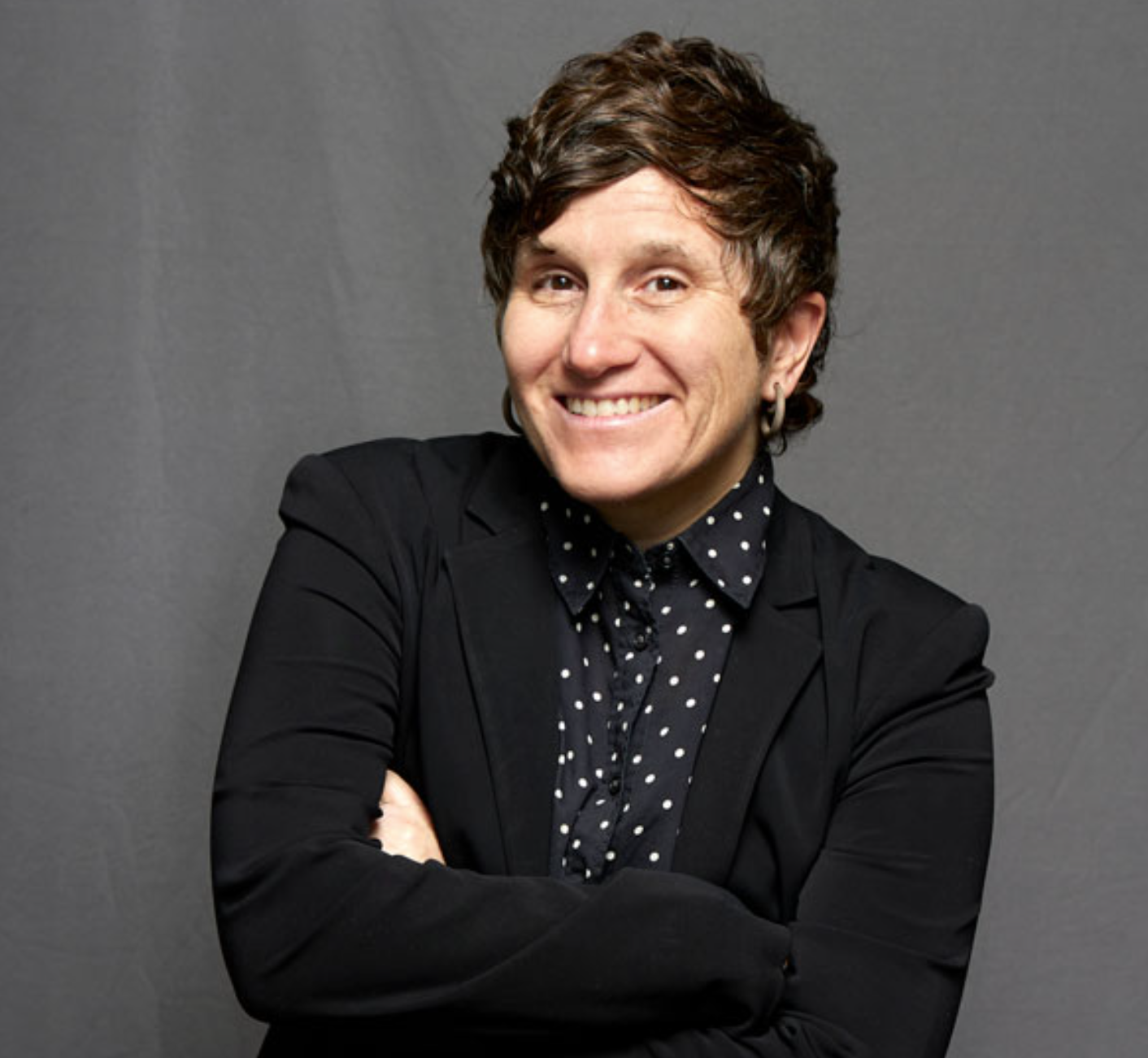 Portrait of a smiling woman with short wavy brown hair, wearing a black blazer and a navy blue polka dot shirt, standing with arms crossed against a gray background.