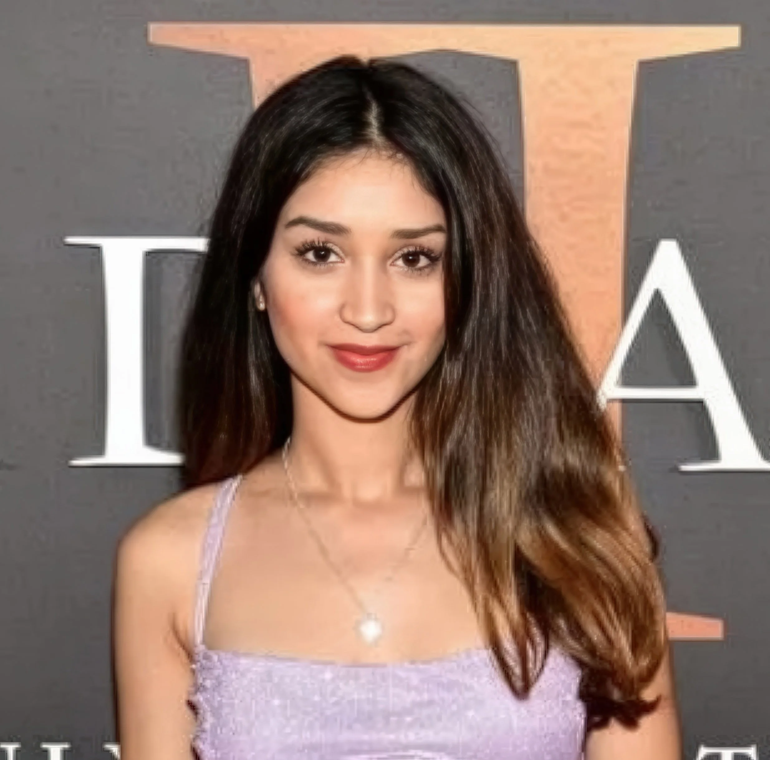 Young woman with long brown hair wearing a lavender spaghetti strap top and a necklace, standing in front of a dark background with partially visible large white and orange letters.