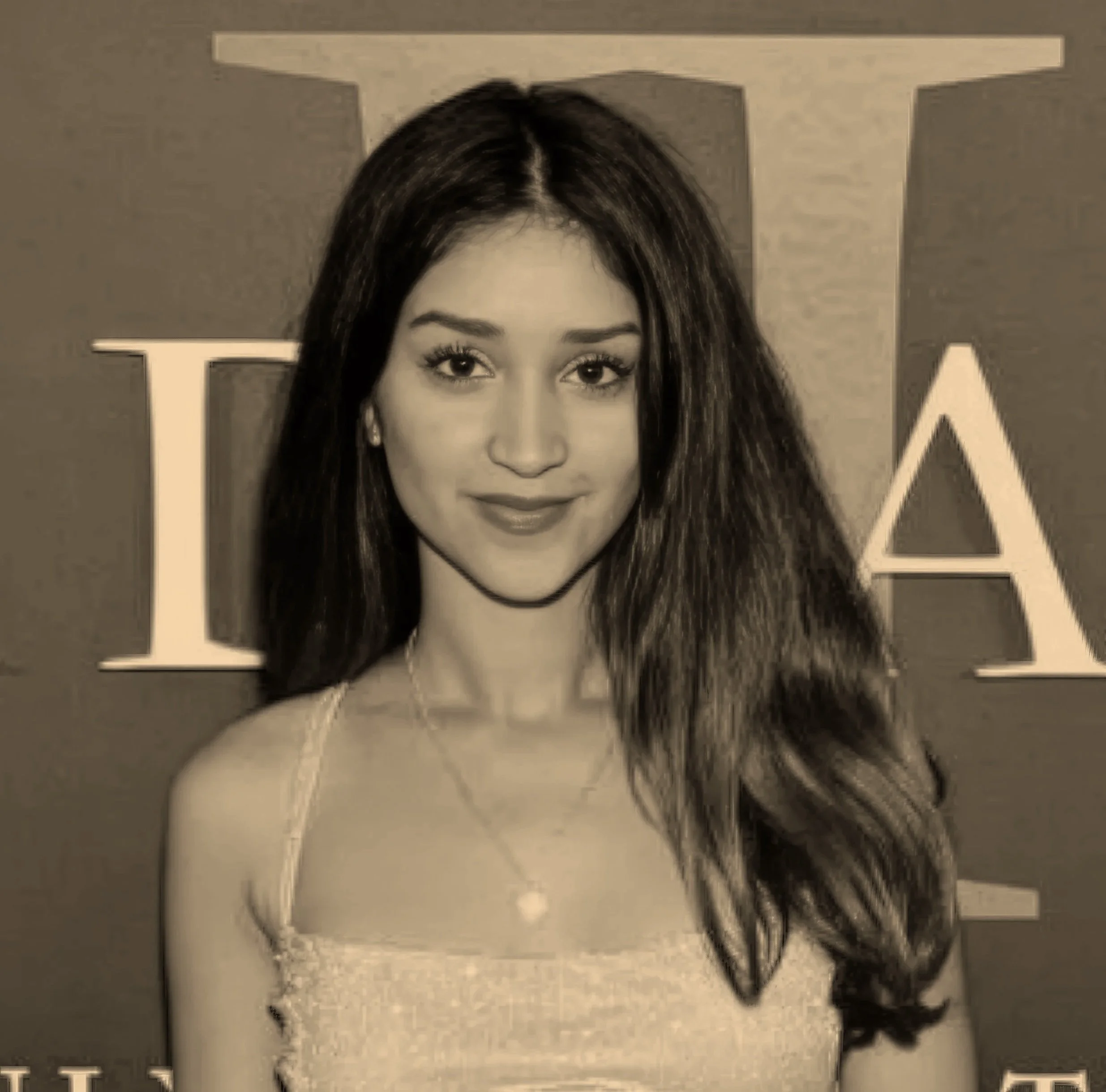 A young woman with long dark hair and a friendly smile posing in front of a backdrop with partially visible letters.
